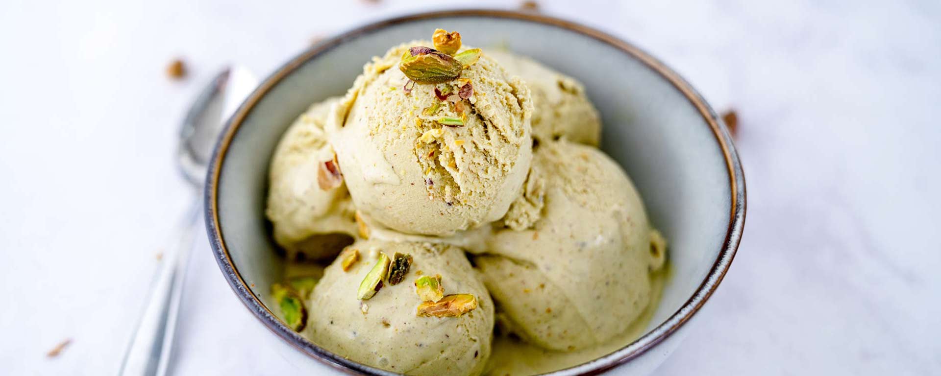 Eine Schüssel mit drei Kugeln Pistazieneis, garniert mit gehackten Pistazien, steht auf einer weißen Fläche, daneben liegt ein Löffel.