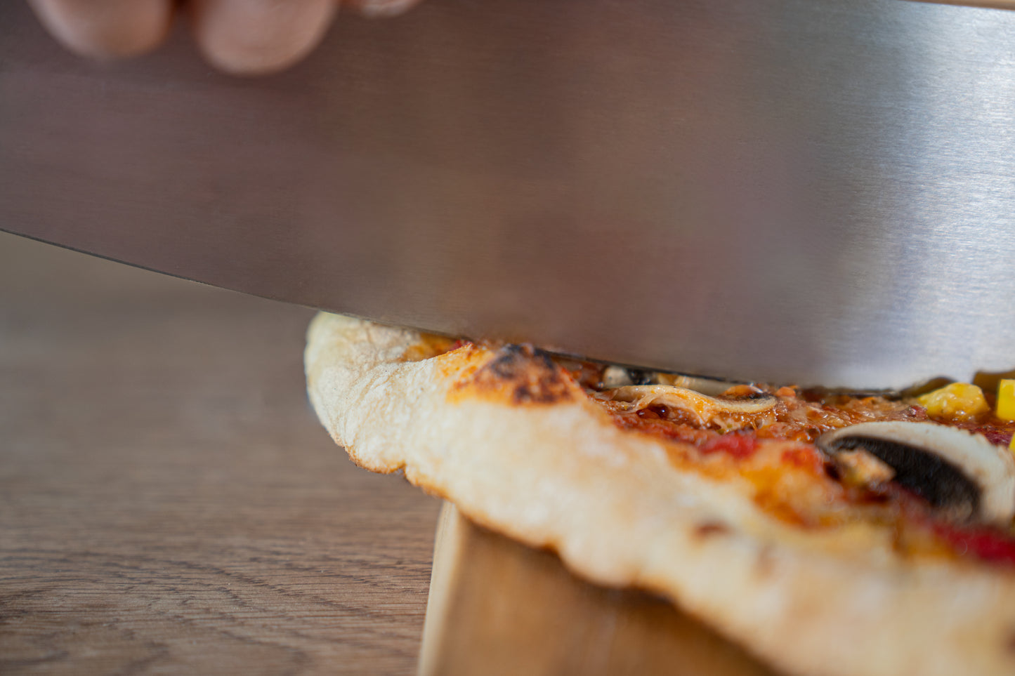 Eine Nahaufnahme einer Pizza, die mit dem Unold Profi Pizzaschneider mit Schneideschutz auf einer Holzunterlage in Stücke geschnitten wird.