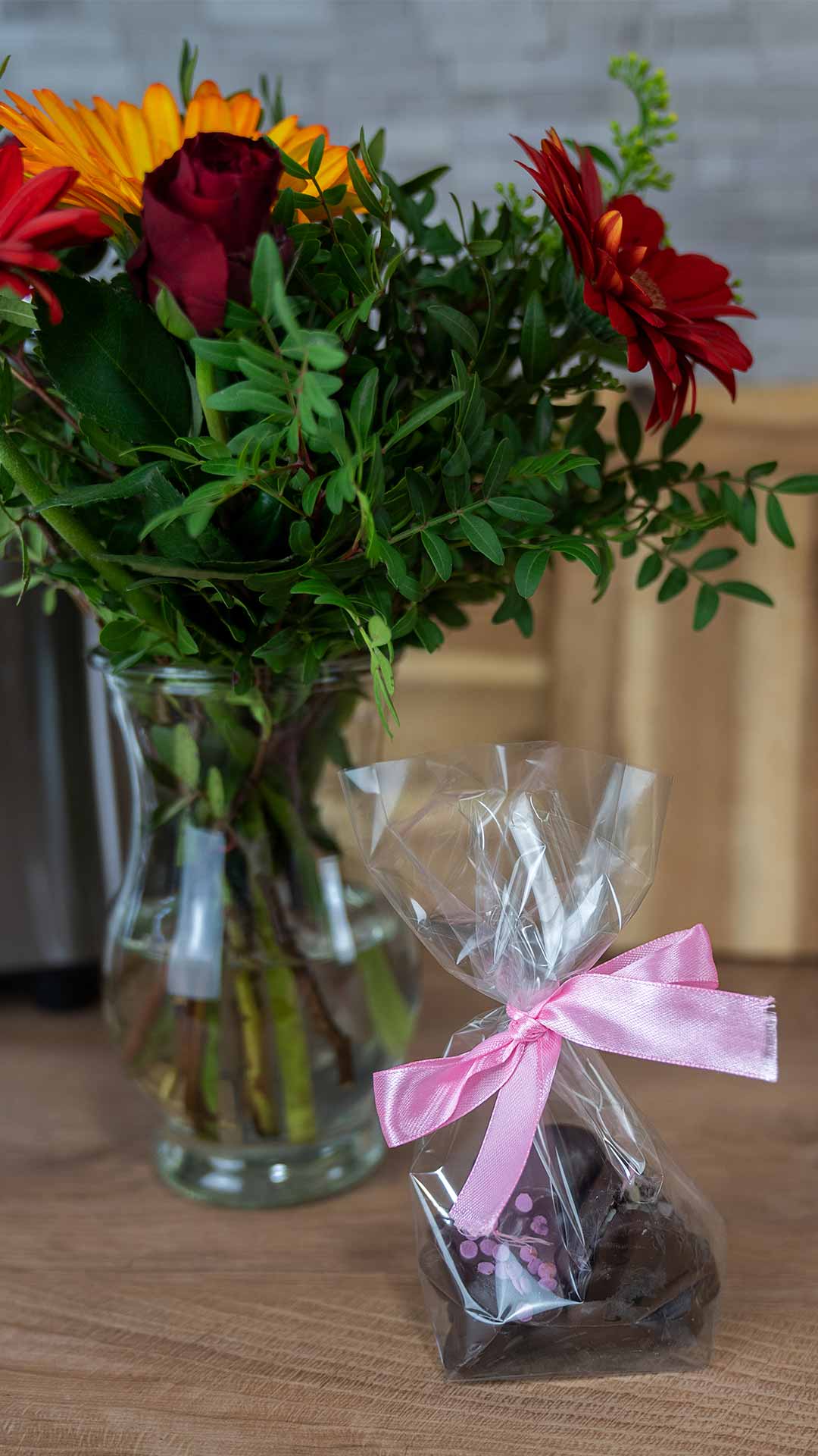 Ein Strauß bunter Blumen in einer Glasvase steht auf einer Holzoberfläche neben einer kleinen Tüte Pralinen, die mit einer rosa Schleife zusammengebunden ist.