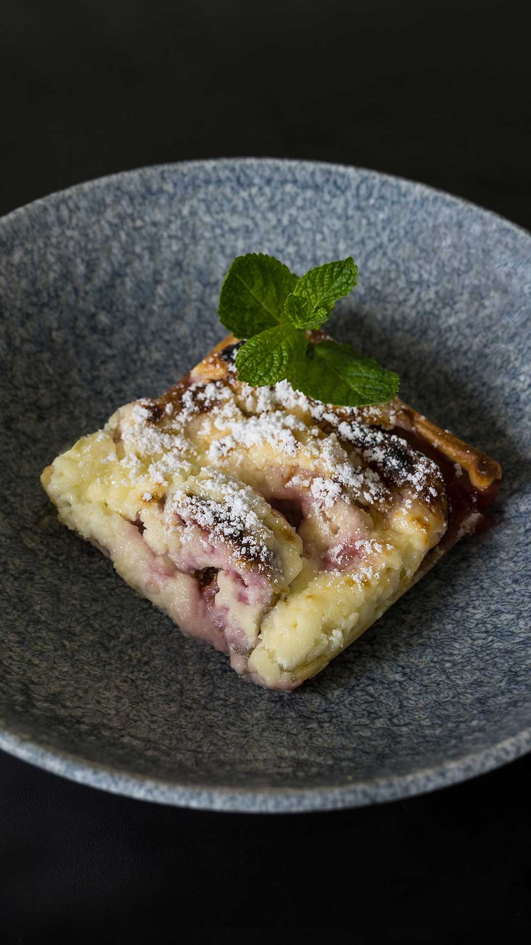 Ein Stück Beeren-Käsekuchen mit Puderzucker und einem Zweig Minze, serviert auf einem strukturierten blaugrauen Teller vor dunklem Hintergrund.