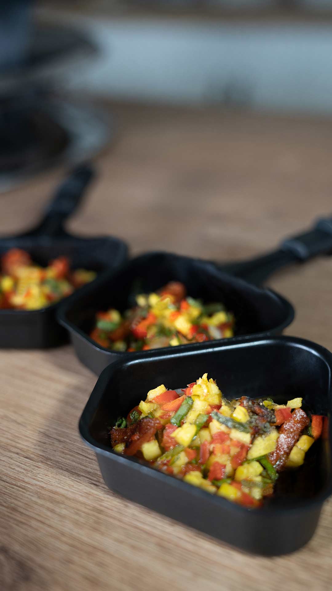 Drei kleine schwarze Servierpfannen, jede mit einer bunten Mischung aus fein geschnittenem Gemüse, darunter gelbe, grüne und rote Stücke, auf einer Holzoberfläche platziert.