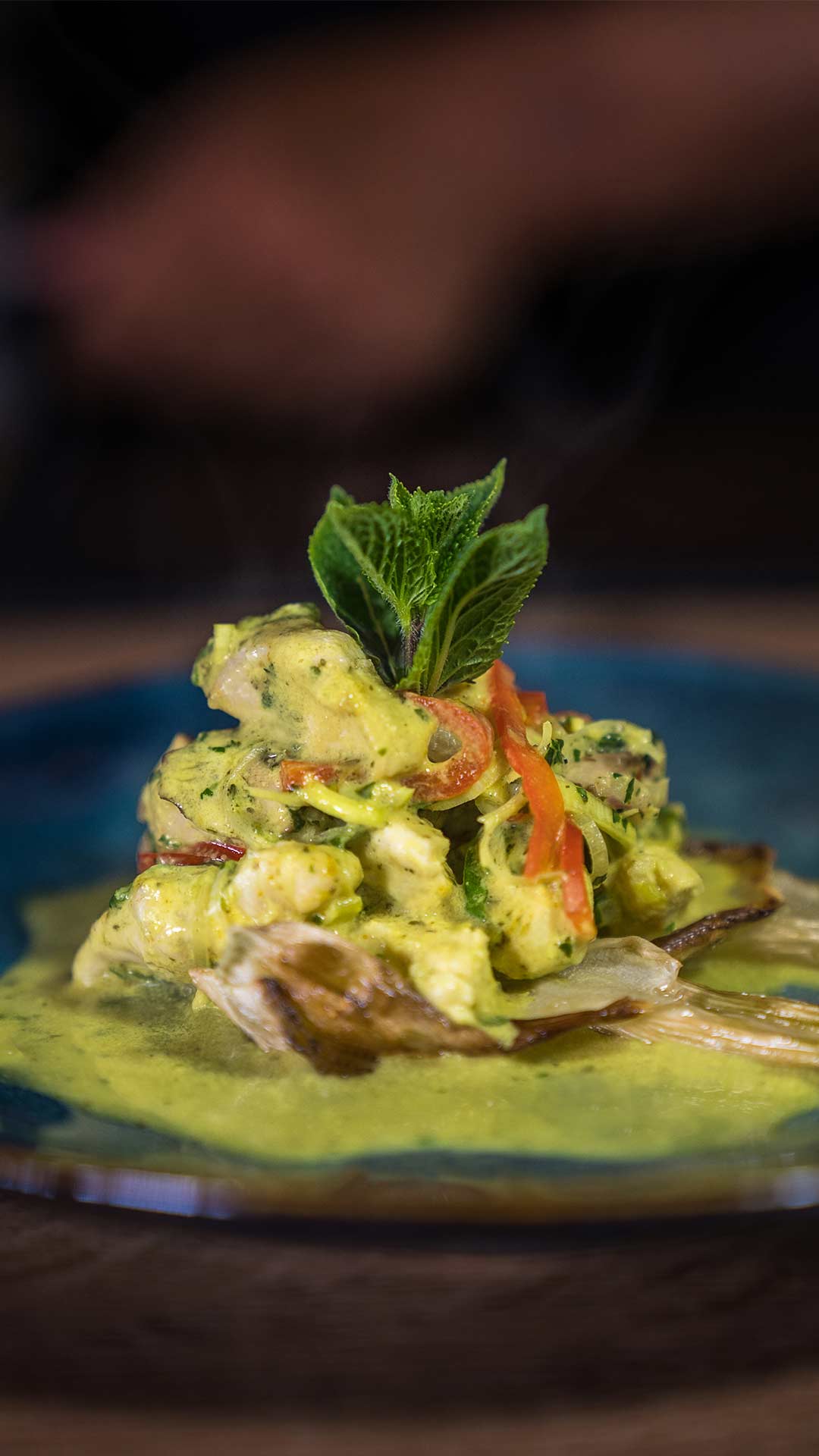 Eine Nahaufnahme eines Gourmetgerichts mit Fischstücken in einer cremigen gelbgrünen Sauce, garniert mit roten Paprikastreifen und frischen Minzblättern, serviert auf einem blauen Teller.