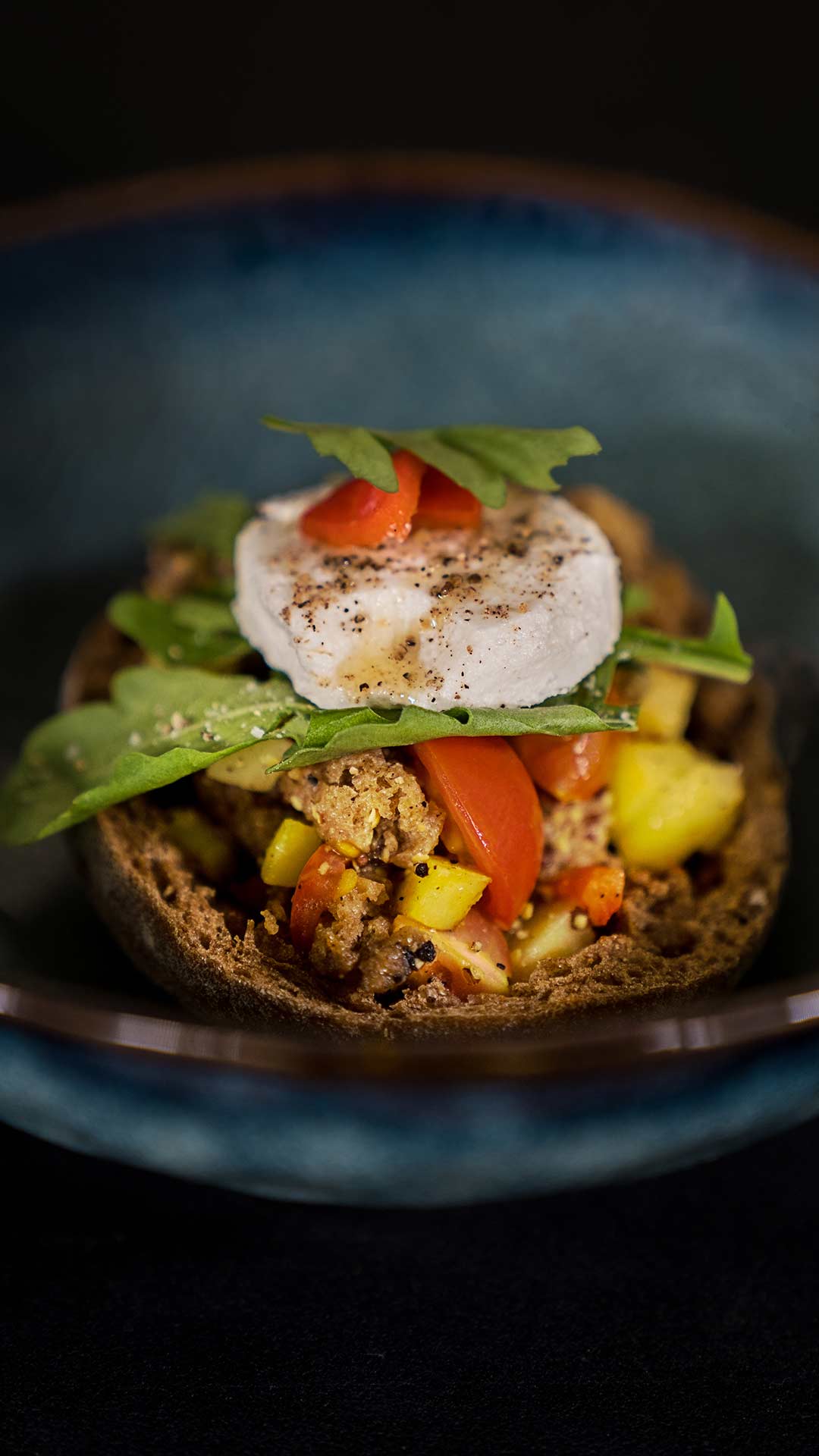 Eine Nahaufnahme eines belegten Brotes mit Blattgemüse, Tomatenscheiben, gewürfelter gelber Paprika, zerbröselter Wurst und einem runden Stück gewürztem Käse, alles auf rustikalem Schwarzbrot in einer blauen Schüssel.