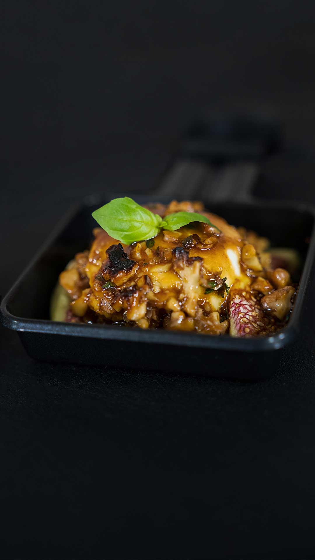 Eine Nahaufnahme einer kleinen gusseisernen Schüssel mit einem herzhaften Überbackengericht, überbacken mit geschmolzenem Käse und einer kräftigen Soße und garniert mit frischen Basilikumblättern, vor einem dunklen Hintergrund.