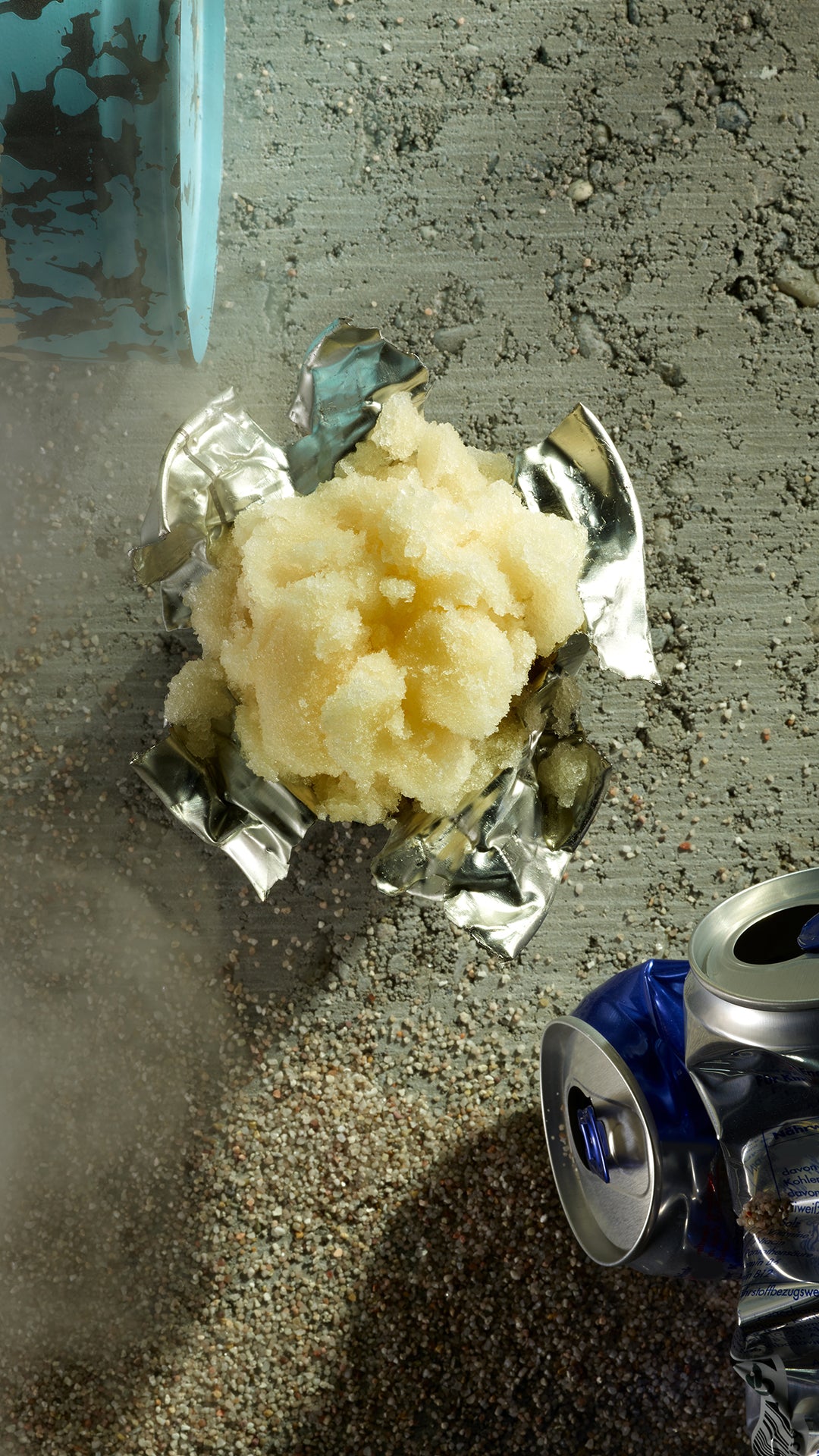 Eine Portion helles, eisiges Granita steht auf einer zerknitterten, geöffneten Aluminiumdose auf einem strukturierten, sandigen Untergrund. Im Hintergrund sind eine weitere zerdrückte Dose und ein blauer Becher teilweise sichtbar.