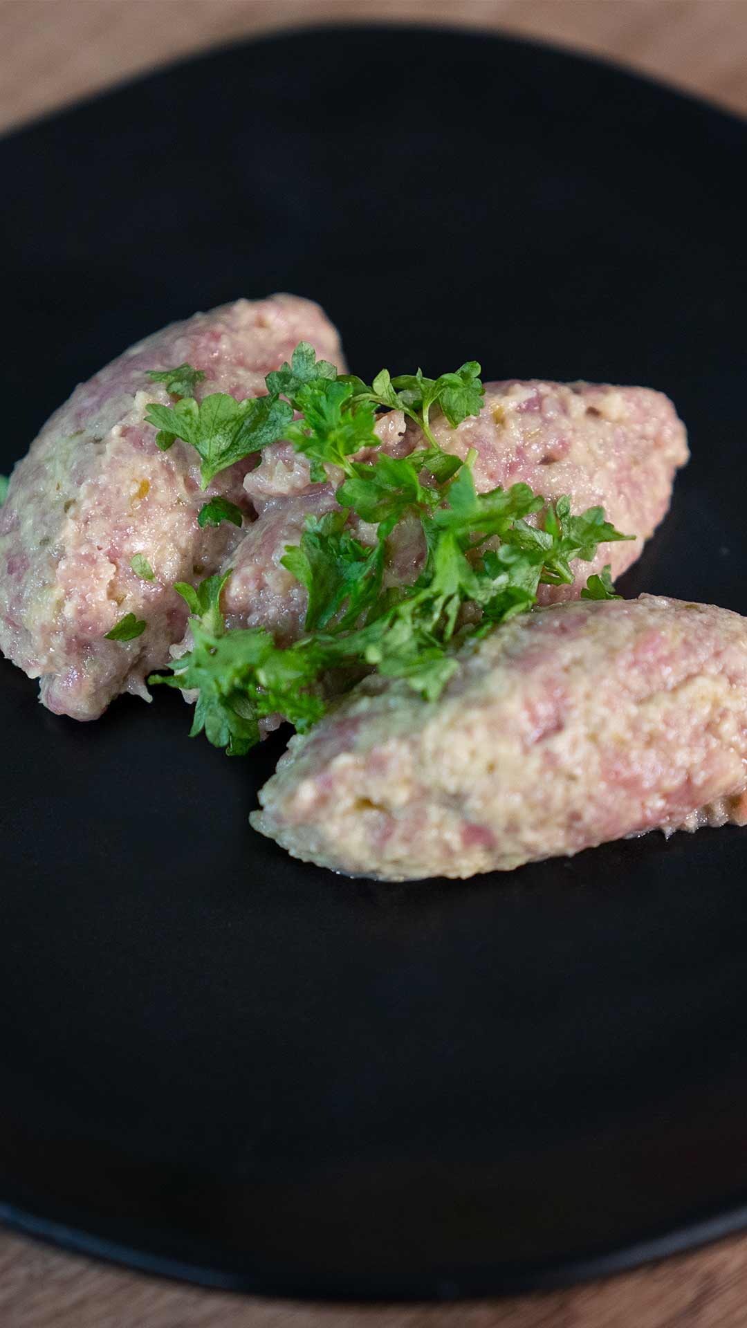 Drei ovale Portionen einer Hackfleisch-Getreide-Mischung werden mit frischer Petersilie garniert auf einem schwarzen Teller serviert.
