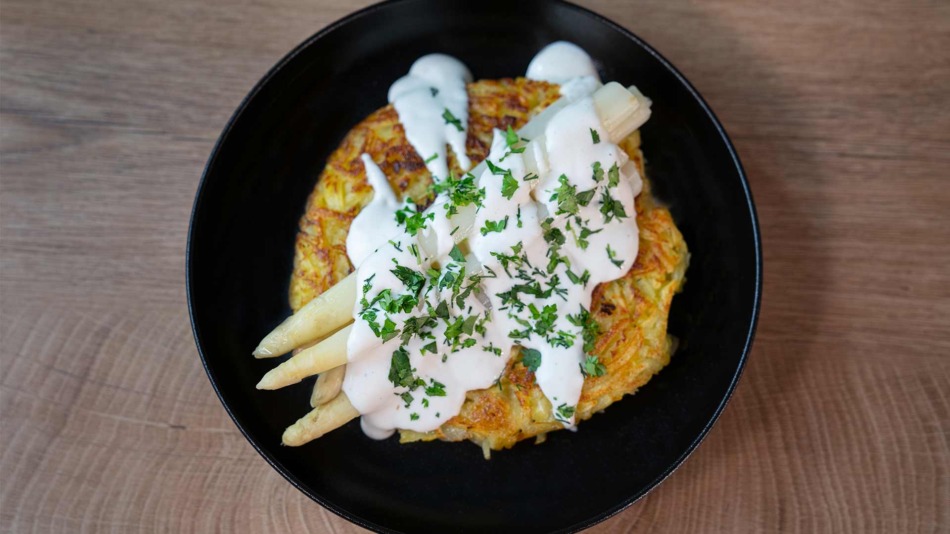 Eine schwarze Schüssel mit knusprigem Kartoffelrösti, garniert mit weißen Spargelstangen, cremiger weißer Sauce und gehackter frischer Petersilie, platziert auf einer Holzoberfläche.