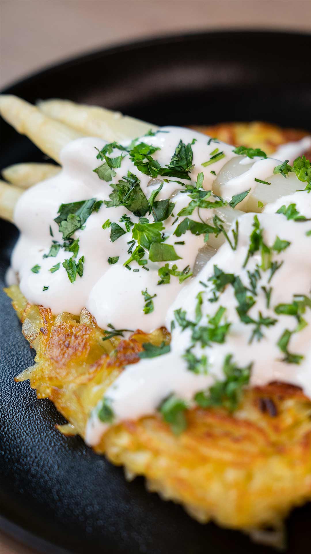 Nahaufnahme einer Kartoffelrösti mit weißem Spargel, cremiger weißer Sauce und gehackten frischen Kräutern, serviert auf einem schwarzen Teller.