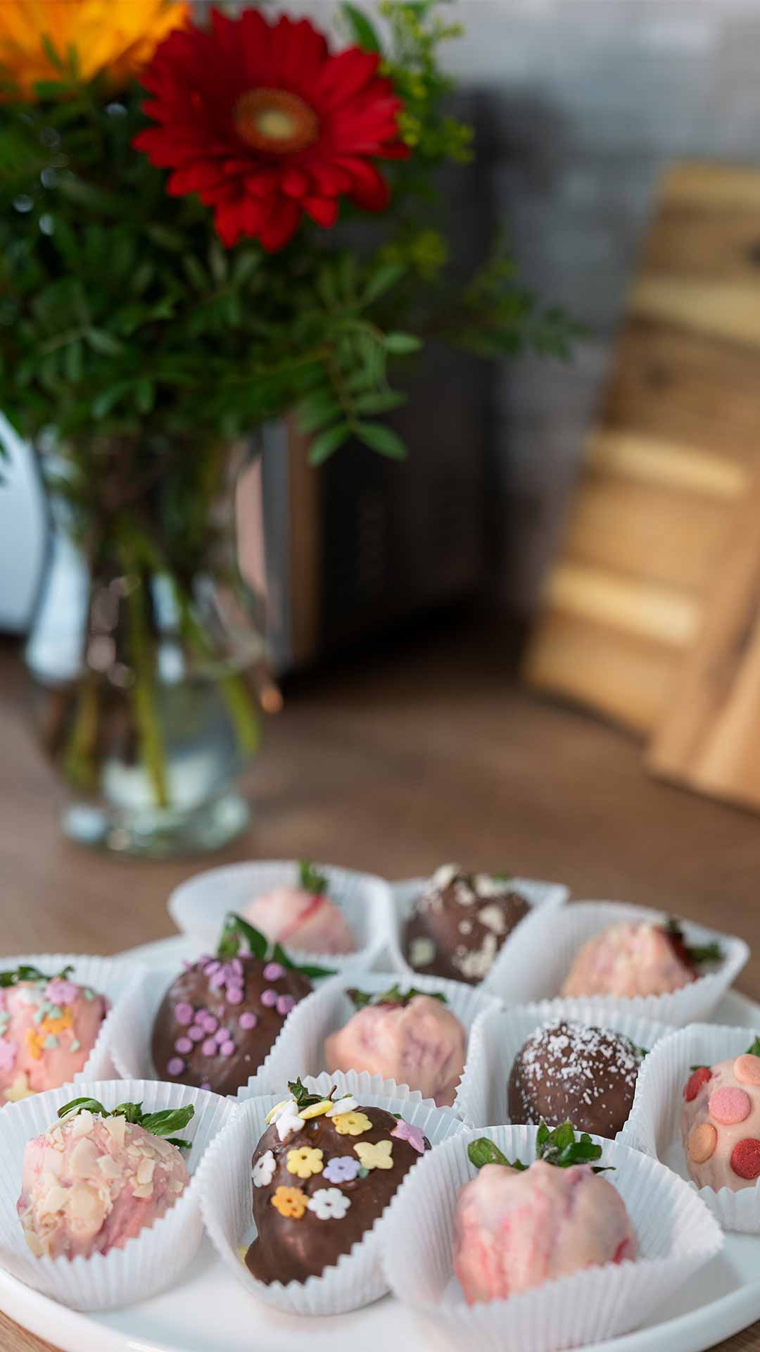 Auf einer Küchentheke steht ein Teller mit dekorierten, schokoladenüberzogenen Erdbeeren in Pappbechern, im Hintergrund eine Vase mit bunten Blumen und ein hölzernes Schneidebrett.