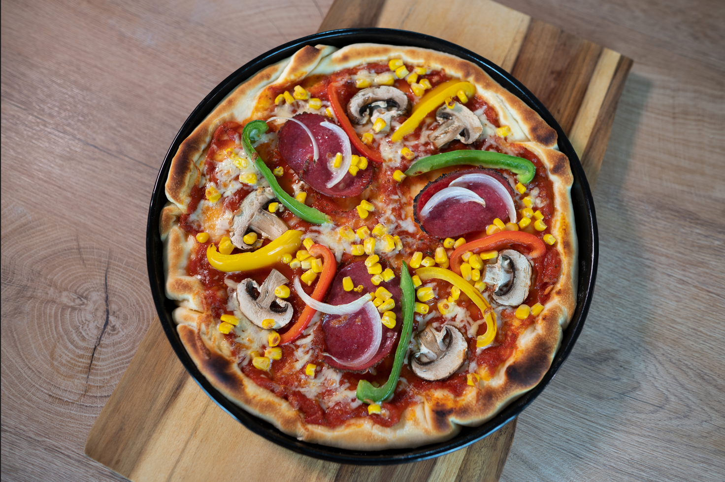 Eine hausgemachte Pizza mit Peperoni, Pilzen, grünen und gelben Paprika, roten Zwiebeln und Mais auf einem schwarzen Blech auf einem Holzbrett.