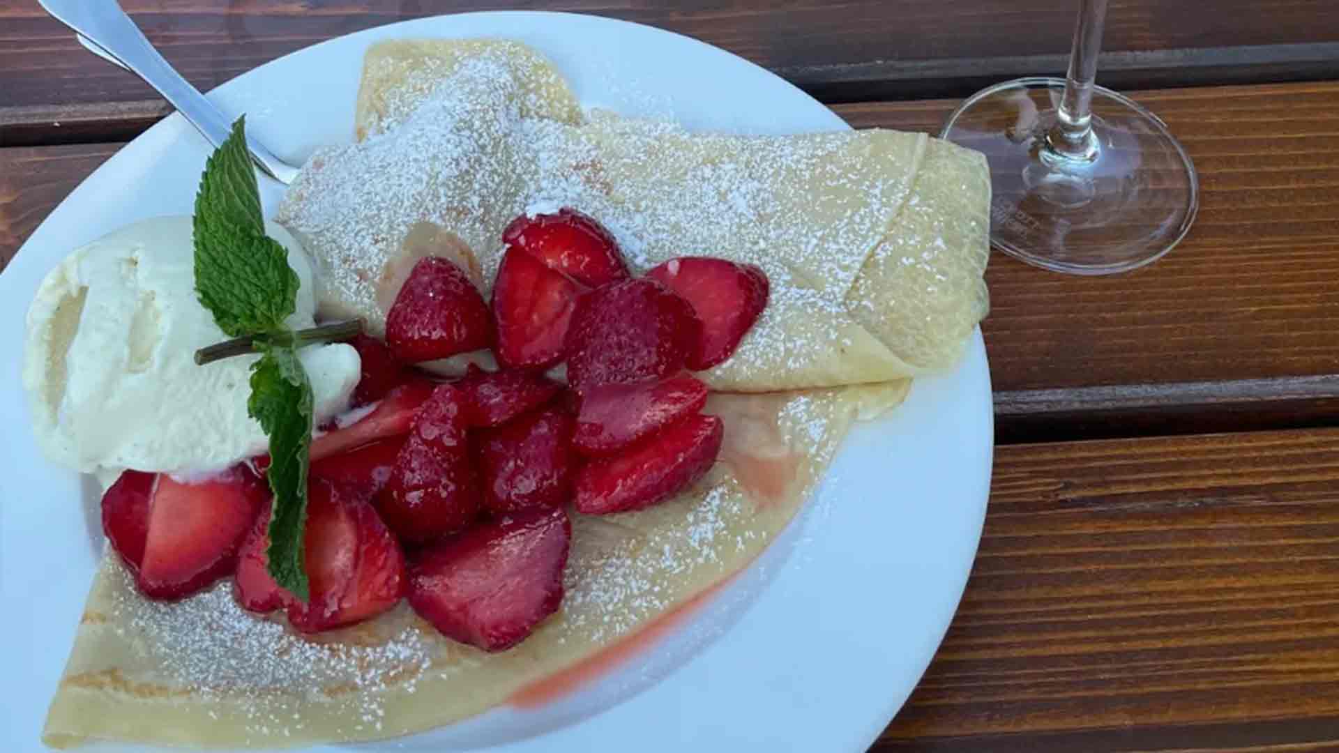 Ein Teller mit einem gefalteten Crêpe, garniert mit Erdbeerscheiben und Puderzucker, serviert mit einer Kugel Vanilleeis und einem Minzzweig, auf einem Holztisch neben einem Stielglas.