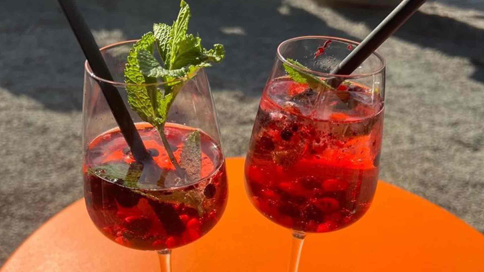 Zwei Gläser eines Cocktails aus roten Beeren, Minzblättern und schwarzen Strohhalmen stehen auf einem orangefarbenen Tisch im Sonnenlicht.