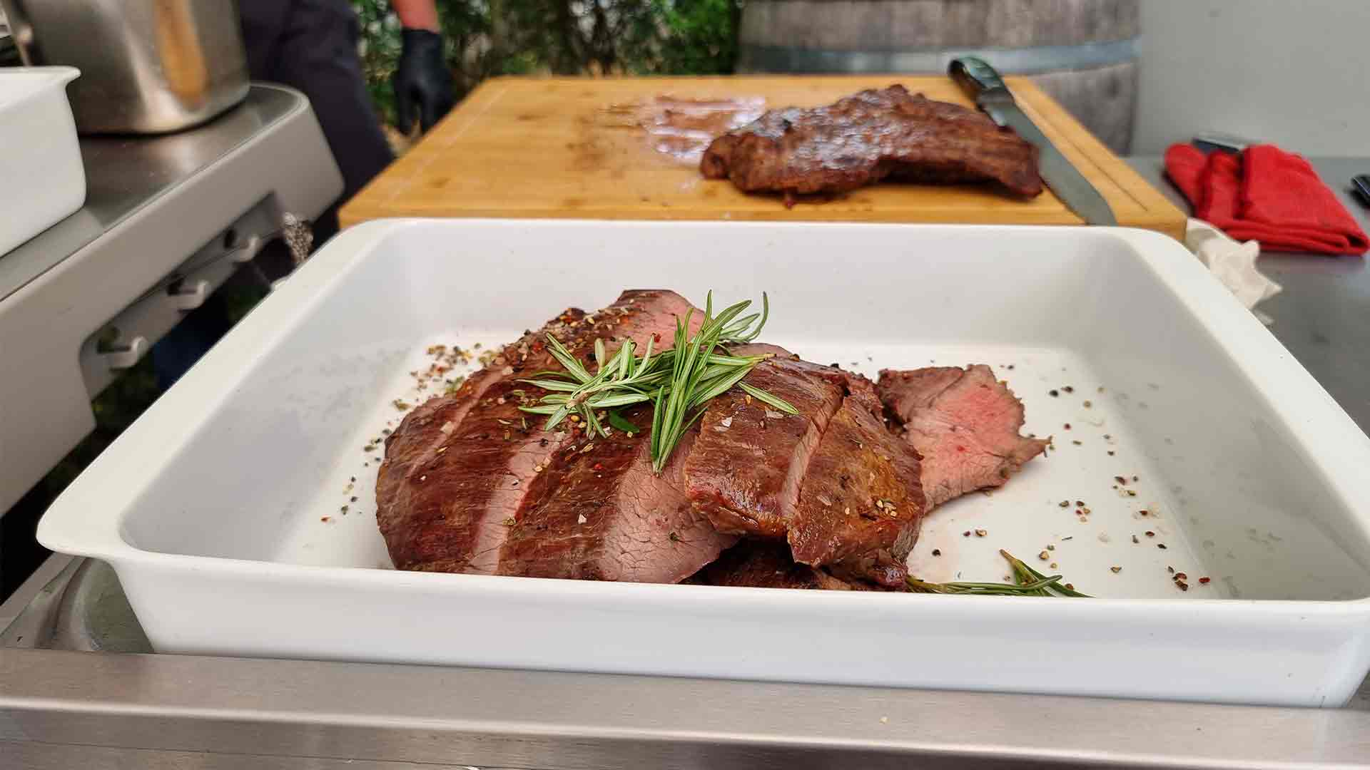 Gegrilltes Steak in Scheiben, garniert mit Rosmarin in einer weißen Schale, mit einem Schneidebrett und noch mehr Steak im Hintergrund auf einer Küchentheke.