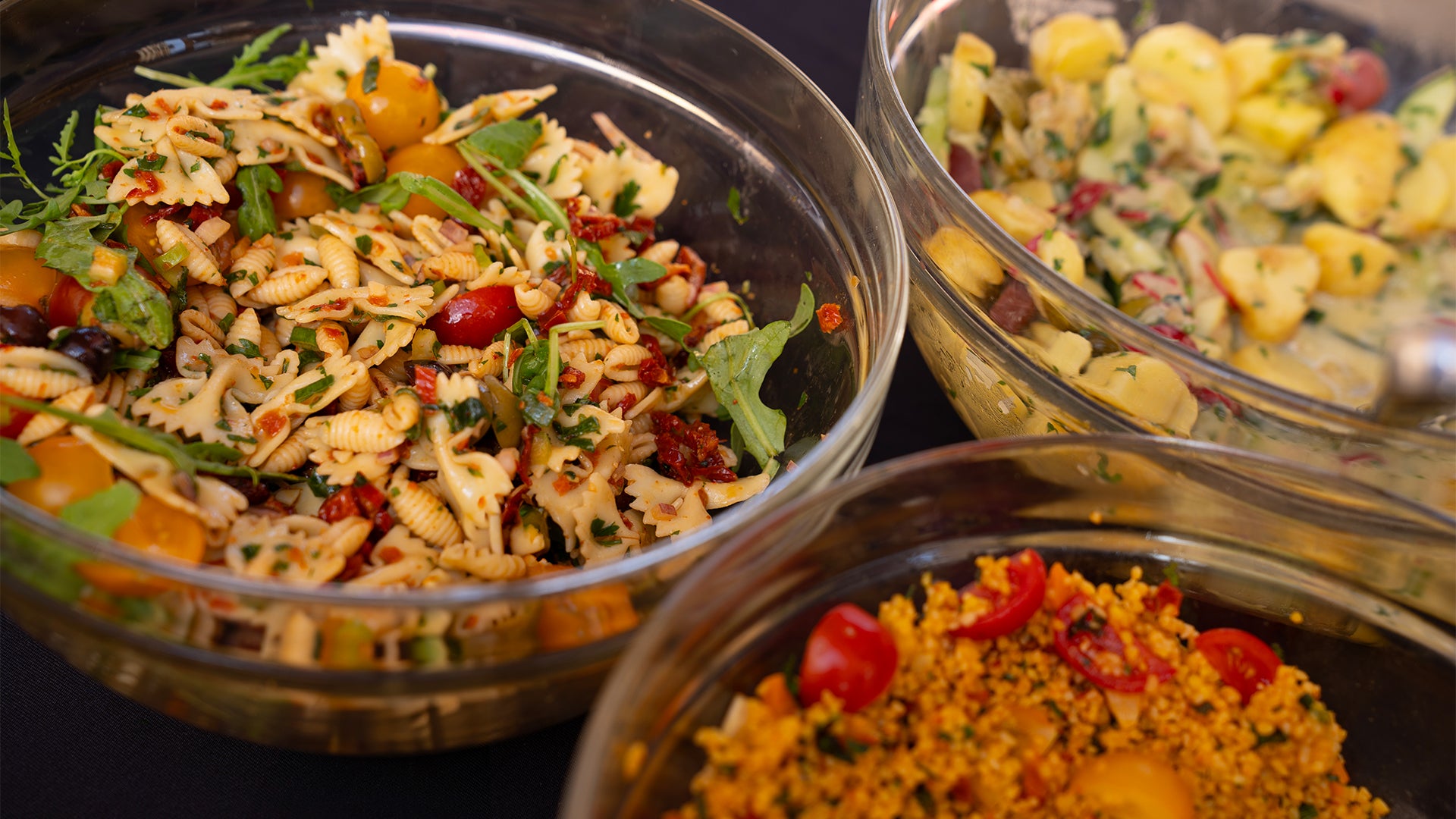 Drei Glasschalen mit bunten Salaten: eine mit Bowtie-Nudeln, Kirschtomaten und Grünzeug, eine weitere mit Kartoffelscheiben und Kräutern und eine dritte mit Couscous, Kirschtomaten und Kräutern.