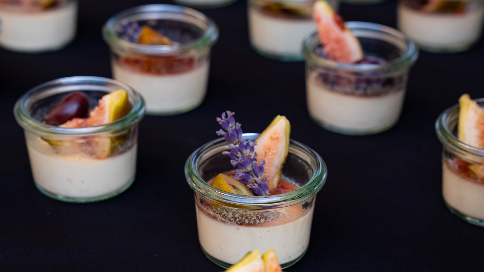 Kleine Gläser, gefüllt mit einem cremigen Dessert, jeweils mit Feigenscheiben, einem Lavendelzweig und anderen Früchten, angeordnet auf einer schwarzen Fläche.