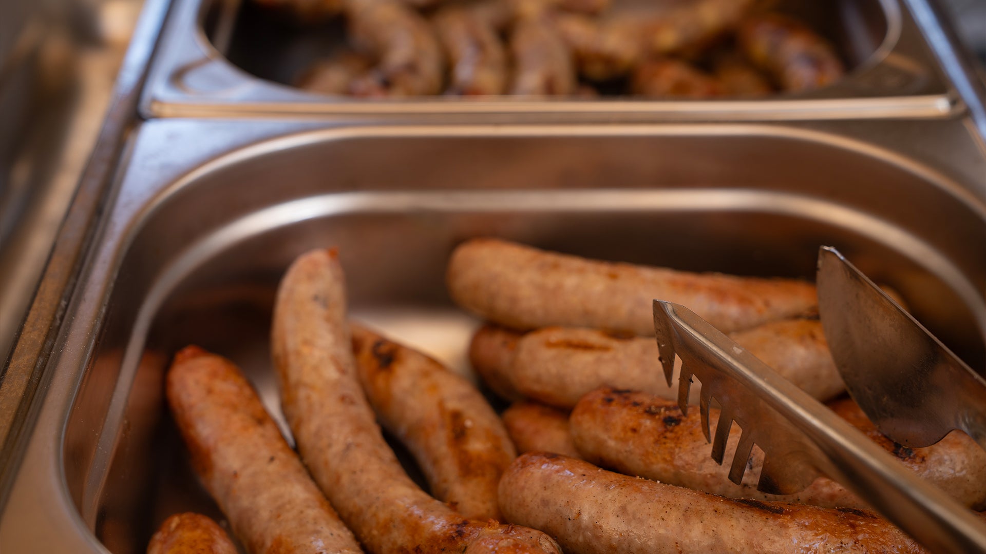 Serviertabletts aus Metall, gefüllt mit gegrillten Würsten, mit einer Metallzange, die auf dem Tablett im Vordergrund ruht. Der Fokus liegt auf den Würsten im vorderen Tablett, weitere Würste sind im Hintergrund zu sehen.