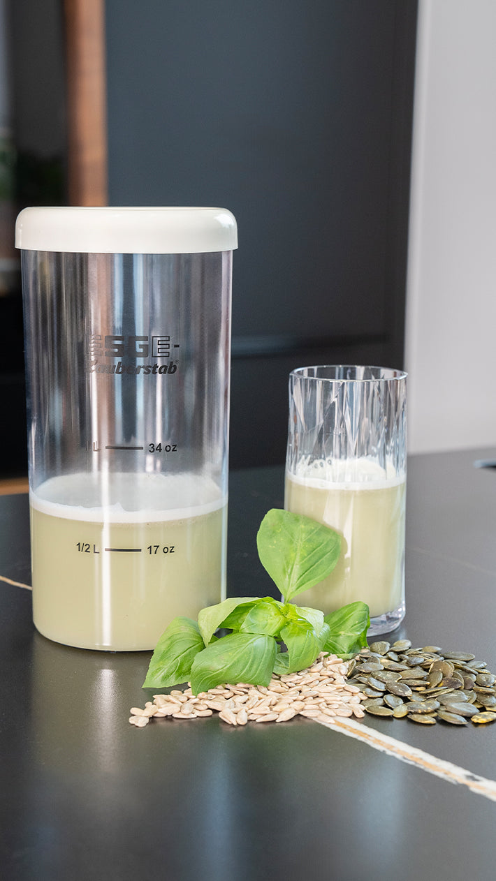 Auf einem dunklen Untergrund stehen ein Glas und ein Behälter mit einem hellgrünen Getränk, daneben grüne Blätter und verstreute Kürbis- und Sonnenblumenkerne.