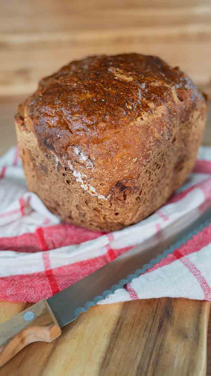 Ein Laib knuspriges Schwarzbrot liegt auf einem rot-weiß karierten Tuch neben einem gezackten Brotmesser auf einer Holzoberfläche.
