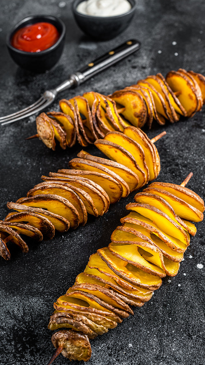 Drei spiralförmig geschnittene Kartoffeln auf Spießen werden auf einer dunklen Oberfläche angerichtet, mit kleinen Schüsseln Ketchup und einem cremigen weißen Dip serviert, mit einer Gabel und einem Messer in der Nähe.