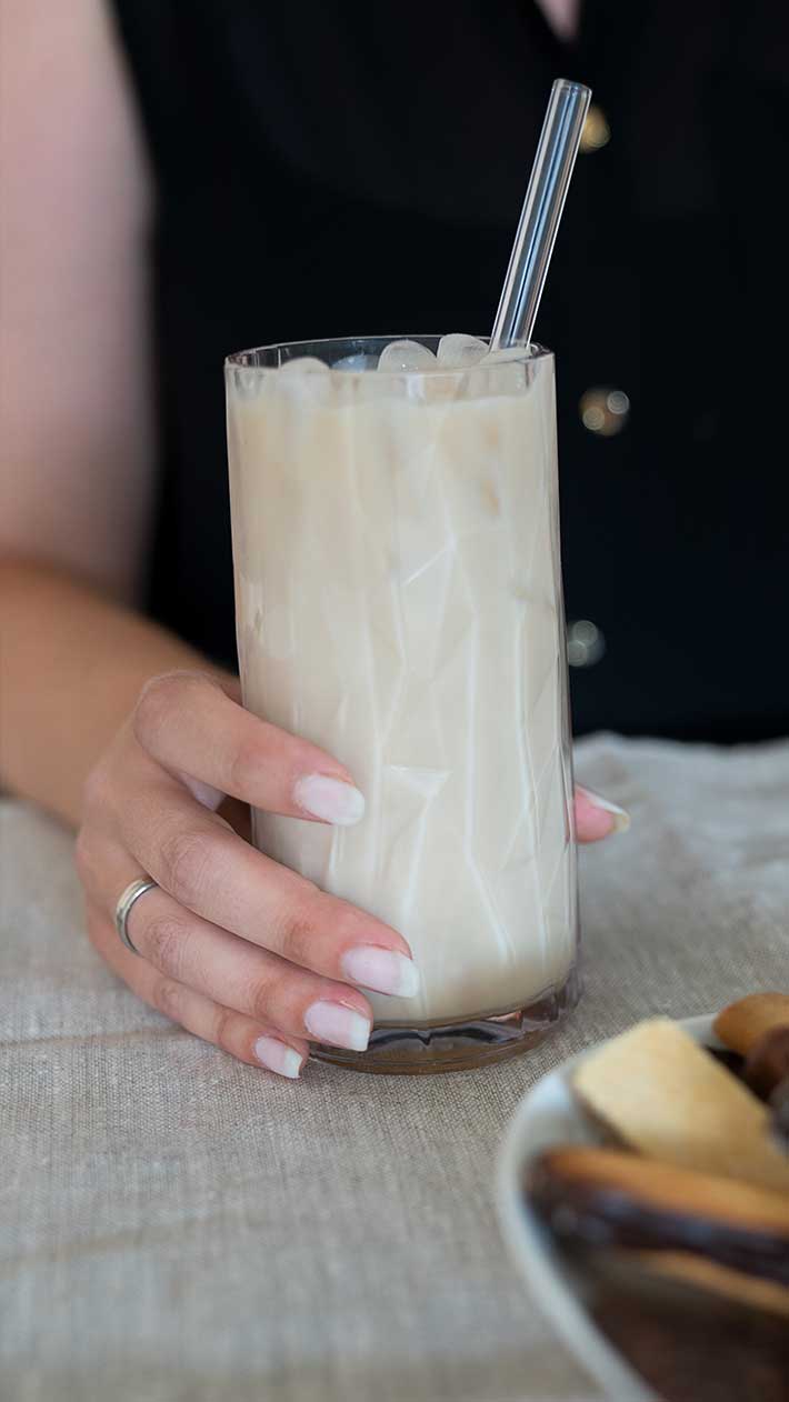 Eine Person mit manikürten Nägeln und einem silbernen Ring hält ein Glas Eistee mit Milch und einem Strohhalm in der Hand. Das Glas steht auf einer beigen Tischdecke neben einem Teller mit verschiedenen Snacks.