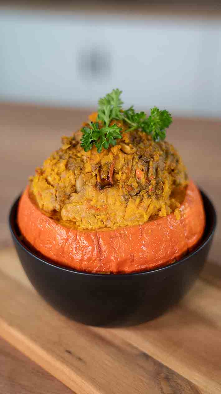 In einer schwarzen Schüssel befindet sich ein gerösteter orangefarbener Kürbis mit einer herzhaften Füllung, garniert mit Petersilie und auf einer Holzoberfläche platziert.