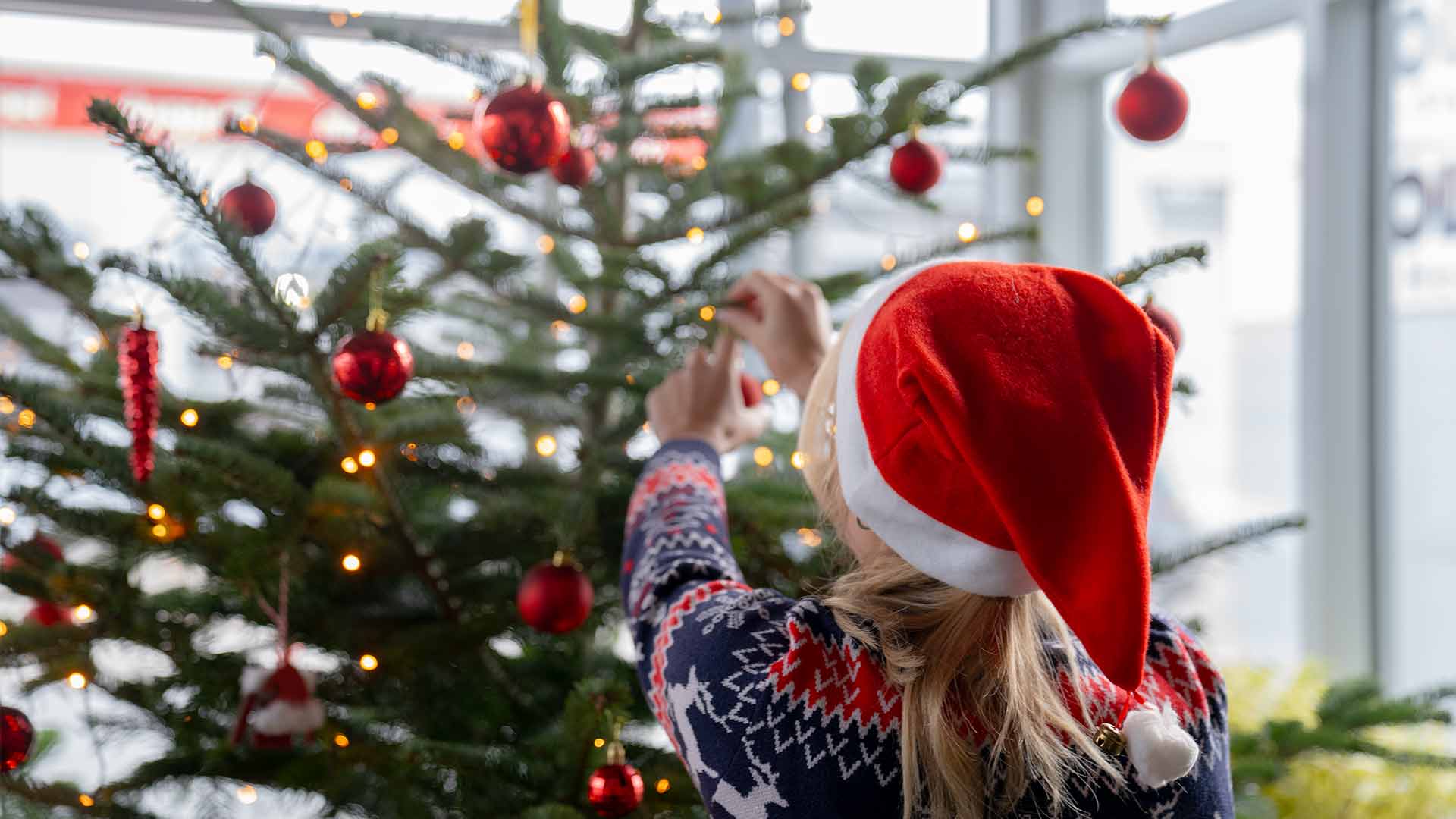 Eine Person mit Weihnachtsmannmütze und festlichem Pullover schmückt in einem hellen Raum einen Weihnachtsbaum mit rotem Schmuck und Lichterketten.