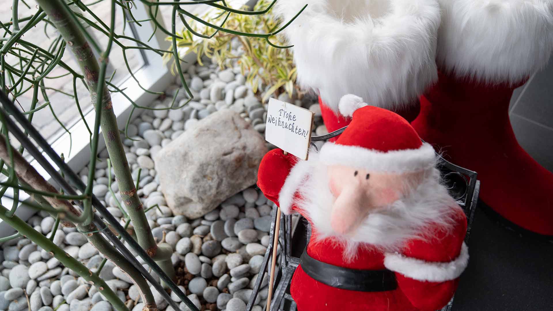 Eine Plüschpuppe des Weihnachtsmanns hält neben großen dekorativen roten Stiefeln ein Schild mit der Aufschrift „Frohe Weihnachten“, umgeben von Kieselsteinen und Pflanzen in der Nähe eines Fensters.