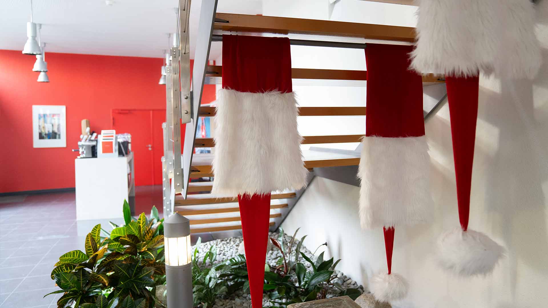Große Weihnachtsmannmützen mit weißem Pelzbesatz hängen an einem modernen Treppengeländer im Innenbereich. Grüne Pflanzen und weiße Steine schmücken den Bereich darunter, im Hintergrund ist eine rote Akzentwand zu sehen.