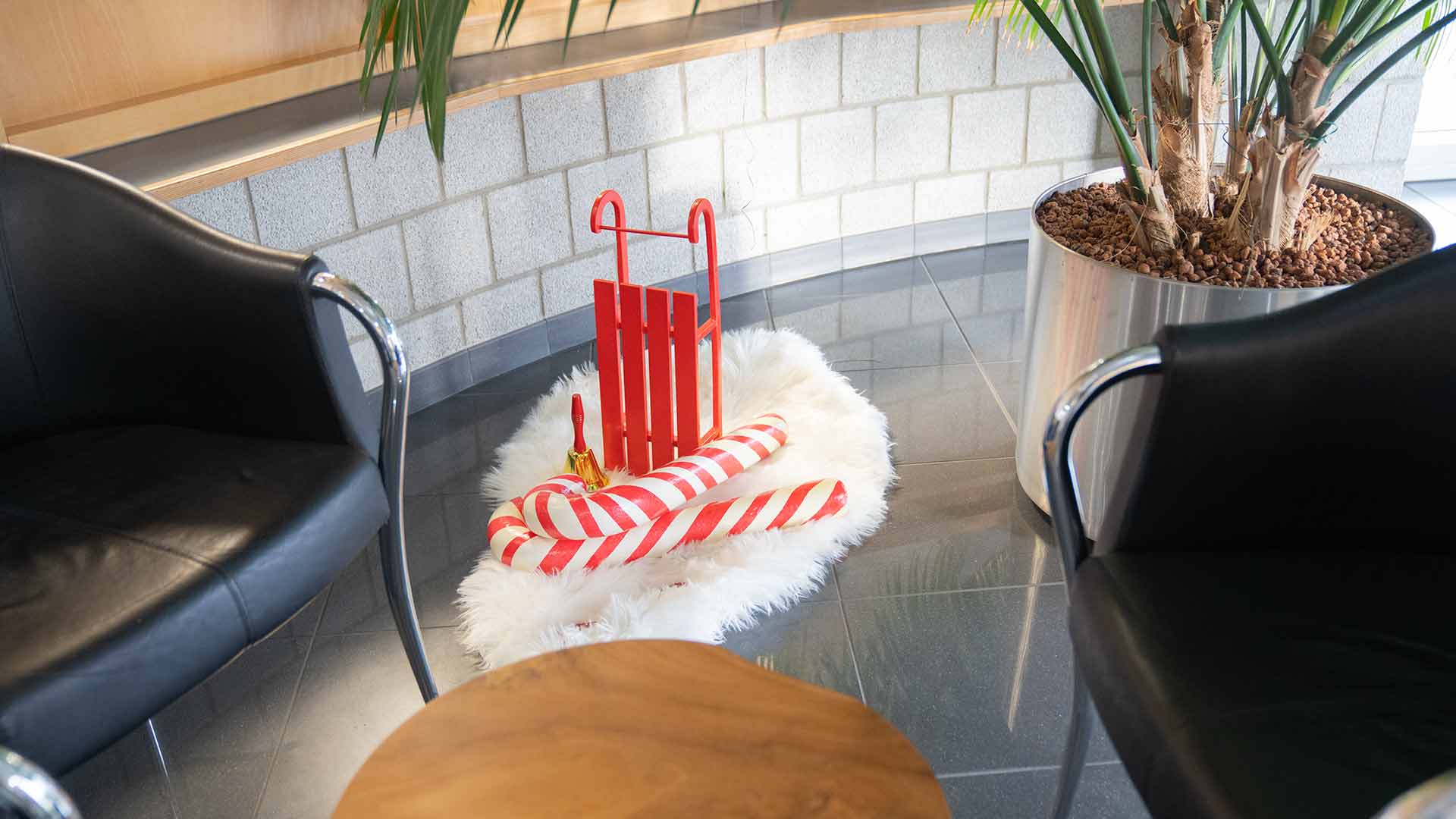 Ein roter Schlitten und zwei übergroße Zuckerstangen liegen auf einem weißen Fellteppich zwischen zwei schwarzen Stühlen in einem modernen Innenraum mit einer großen Topfpflanze.