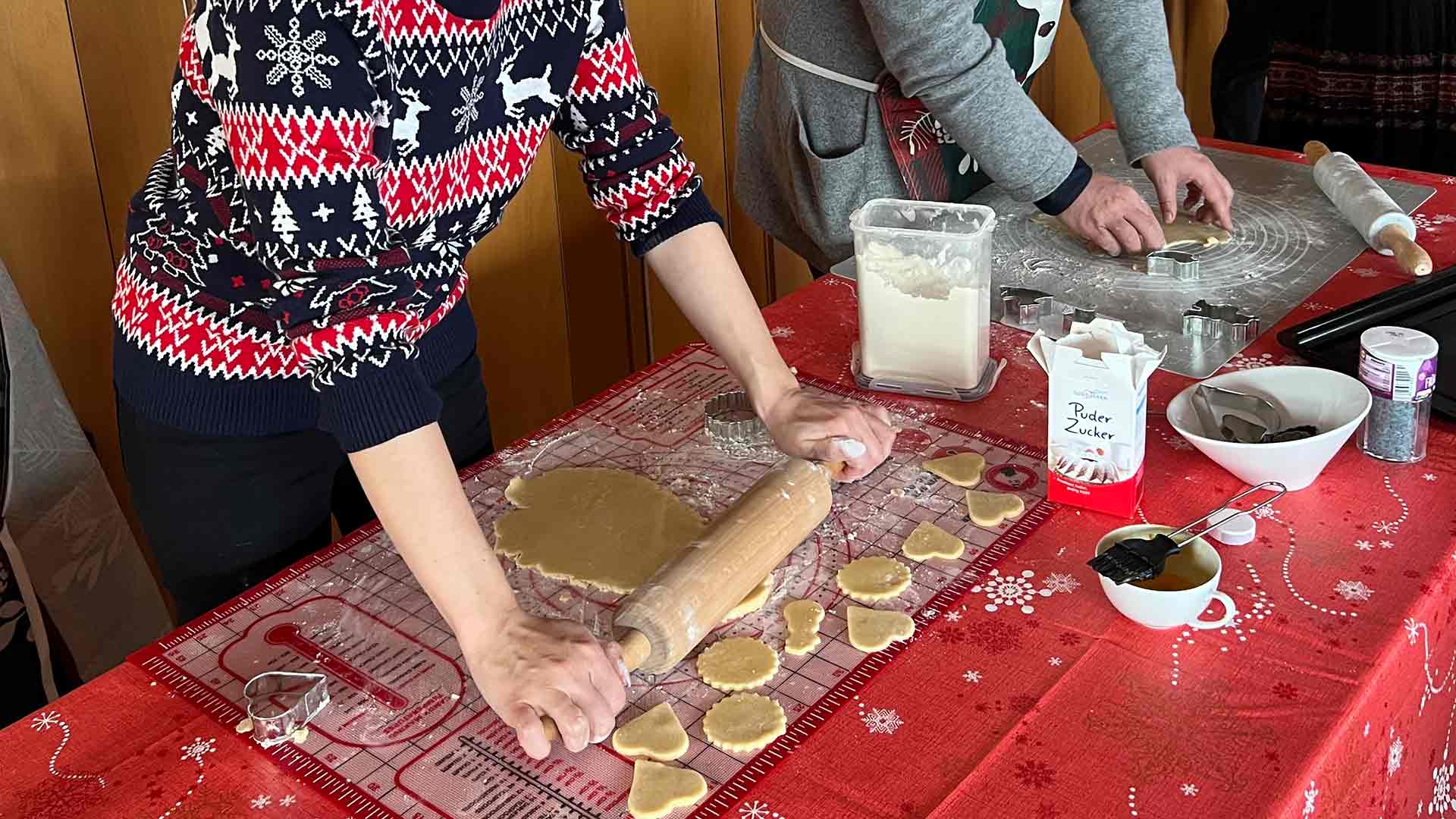 Zwei Personen rollen mit Nudelhölzern Keksteig auf einer roten Tischdecke aus. Verschiedene Ausstechformen, Teigformen und Backzubehör liegen auf dem Tisch verteilt.