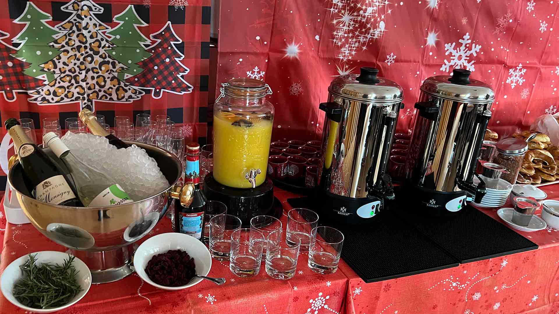 Eine festliche Getränkestation mit zwei großen Kaffeekannen, einem Glasspender mit Orangensaft, einem Eimer mit Weinflaschen auf Eis, verschiedenen Sirups, Gläsern und Weihnachtsdekorationen mit roten und karierten Mustern mit Weihnachtsbäumen.