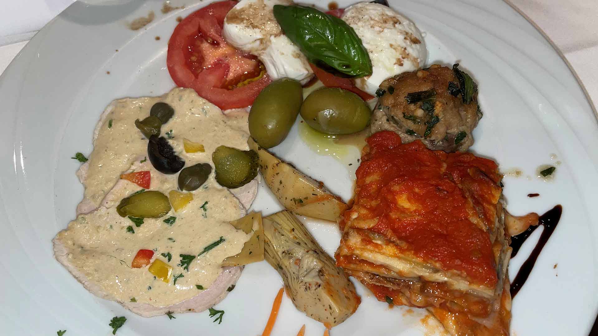 Ein Teller mit verschiedenen italienischen Vorspeisen, darunter geschnittene Tomaten mit Mozzarella und Basilikum, grüne Oliven, Artischockenherzen, Fleischbällchen, Lasagne und Fleischscheiben mit einer Sahnesauce und Kapern.