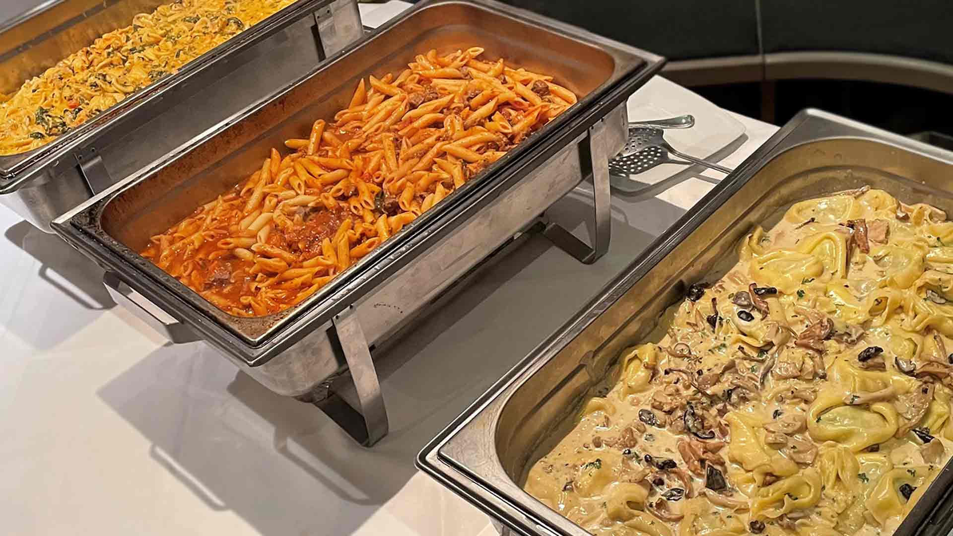 Drei Buffettabletts aus Edelstahl mit verschiedenen Pastagerichten: eines mit Penne in roter Sauce, ein weiteres mit Tortellini in cremiger weißer Sauce mit Pilzen und ein drittes mit Käsepasta. Servierbesteck steht in der Nähe.