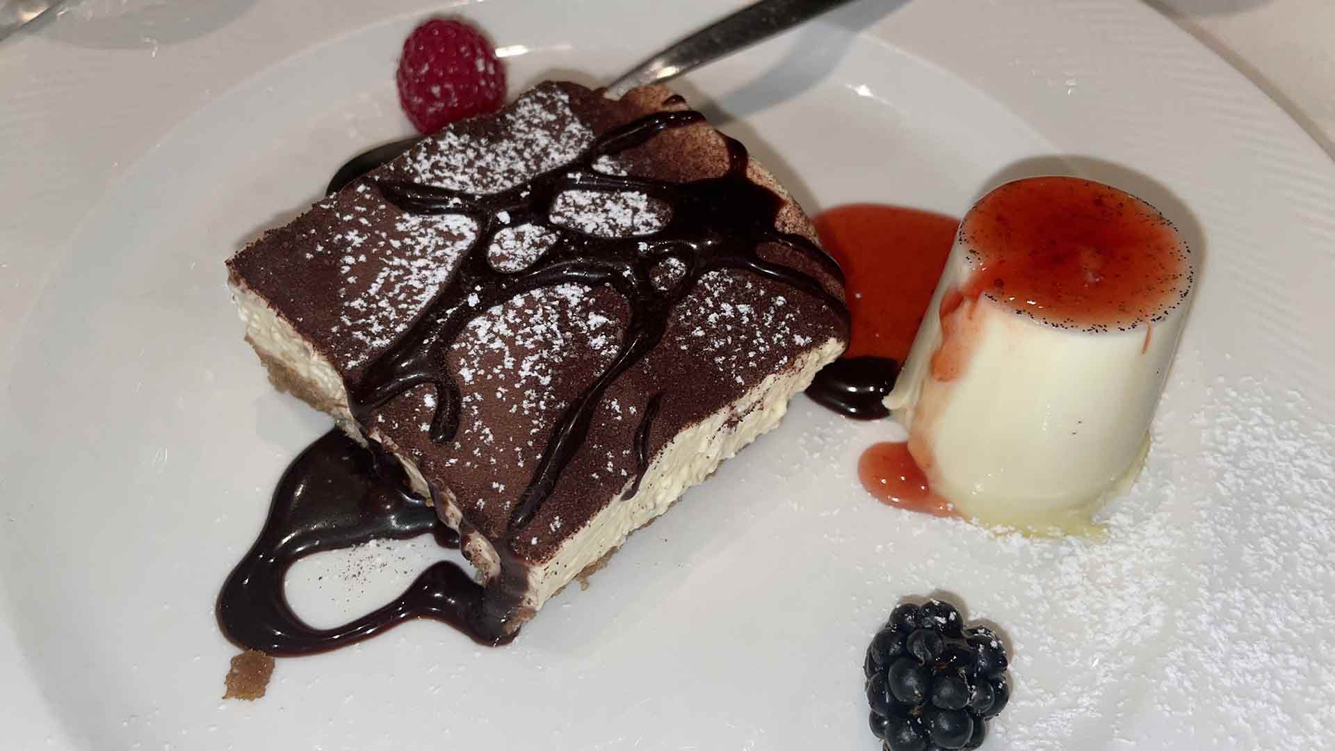 Ein Dessertteller mit einem mit Schokolade beträufelten und mit Puderzucker bestäubten Tiramisu-Quadrat, einer kleinen Panna Cotta mit roter Sauce, einer Himbeere und einer Brombeere auf einem weißen Teller.