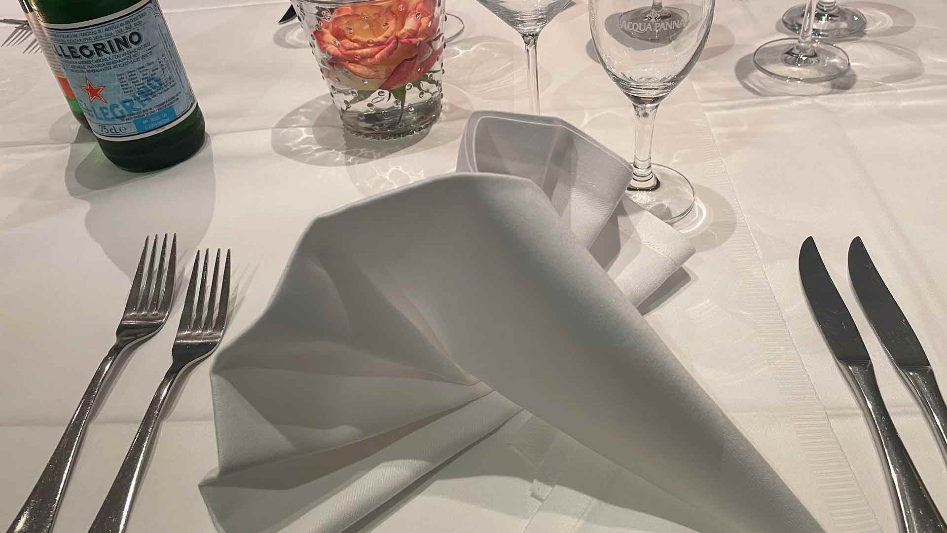 Ein hübsch gedeckter Esstisch mit einer gefalteten weißen Serviette, zwei Gabeln, zwei Messern, klaren Gläsern, einer Flasche San Pellegrino-Sprudelwasser und einer kleinen Glasvase mit einer pfirsichfarbenen Rose.