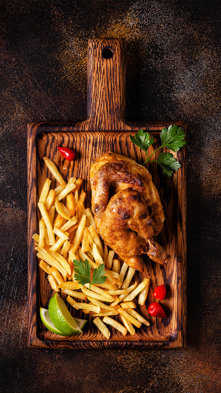 Gegrilltes Hähnchen mit goldbraunen Pommes Frites, Limettenspalten, Kirschtomaten und Petersilie, serviert auf einem Holzschneidebrett vor einem dunklen, strukturierten Hintergrund.