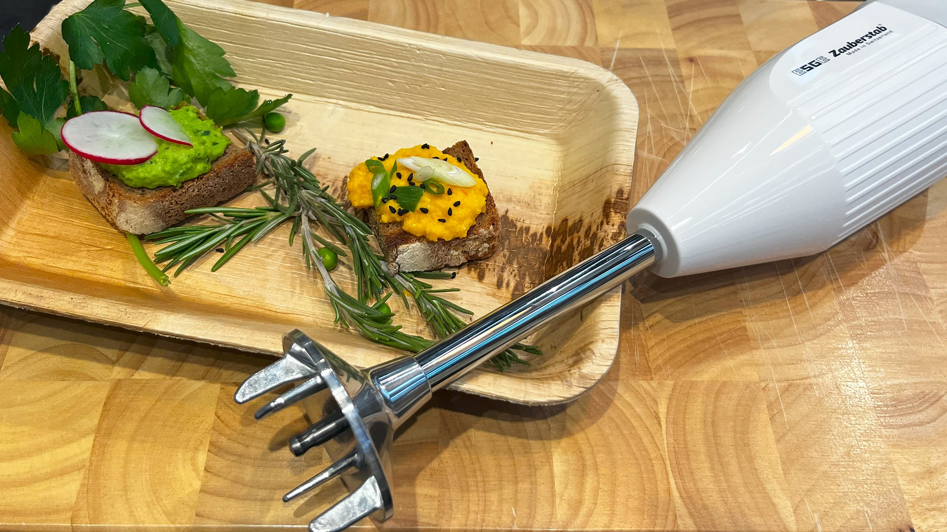 Auf einer Holzfläche liegt ein weißer Stabmixer neben einem Tablett mit zwei Brotscheiben, belegt mit buntem Aufstrich, frischen Kräutern und Radieschenscheiben.