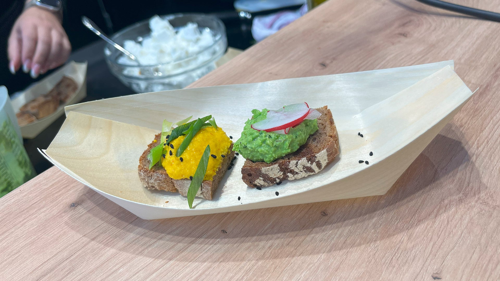 Zwei Brotscheiben auf einer Bambusschale: eine mit gelbem Brotaufstrich und grüner Garnitur, die andere mit grünem Brotaufstrich und Radieschenscheiben, auf einer Holzoberfläche mit einer Schüssel Weißkäse im Hintergrund.