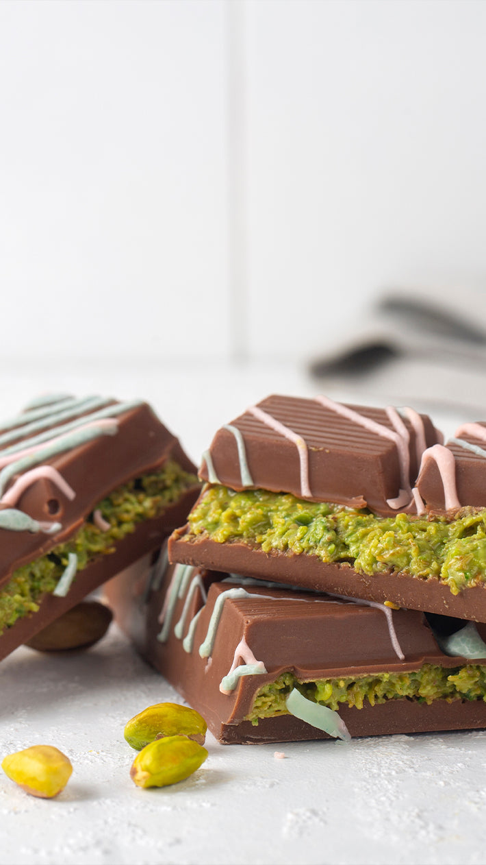 Drei schokoladenüberzogene Pistazienriegel liegen auf einer weißen Fläche, im Inneren ist eine grüne Pistazienfüllung sichtbar. Pistazien und rosa und blaue Zuckerguss-Spritzer zieren die Szene.