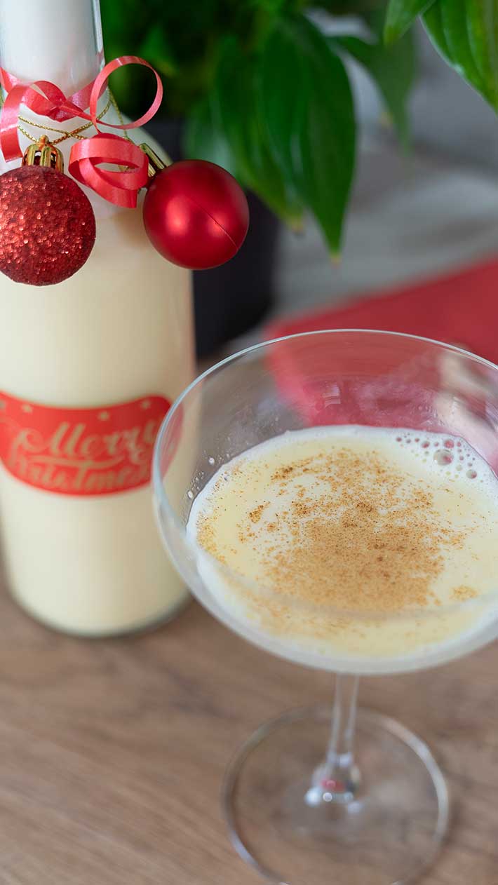 Ein Glas cremiger Eierlikör, bestreut mit Muskatnuss, steht neben einer festlichen Flasche, die mit roten Ornamenten und Bändern geschmückt ist, im Hintergrund sind grüne Blätter zu sehen.