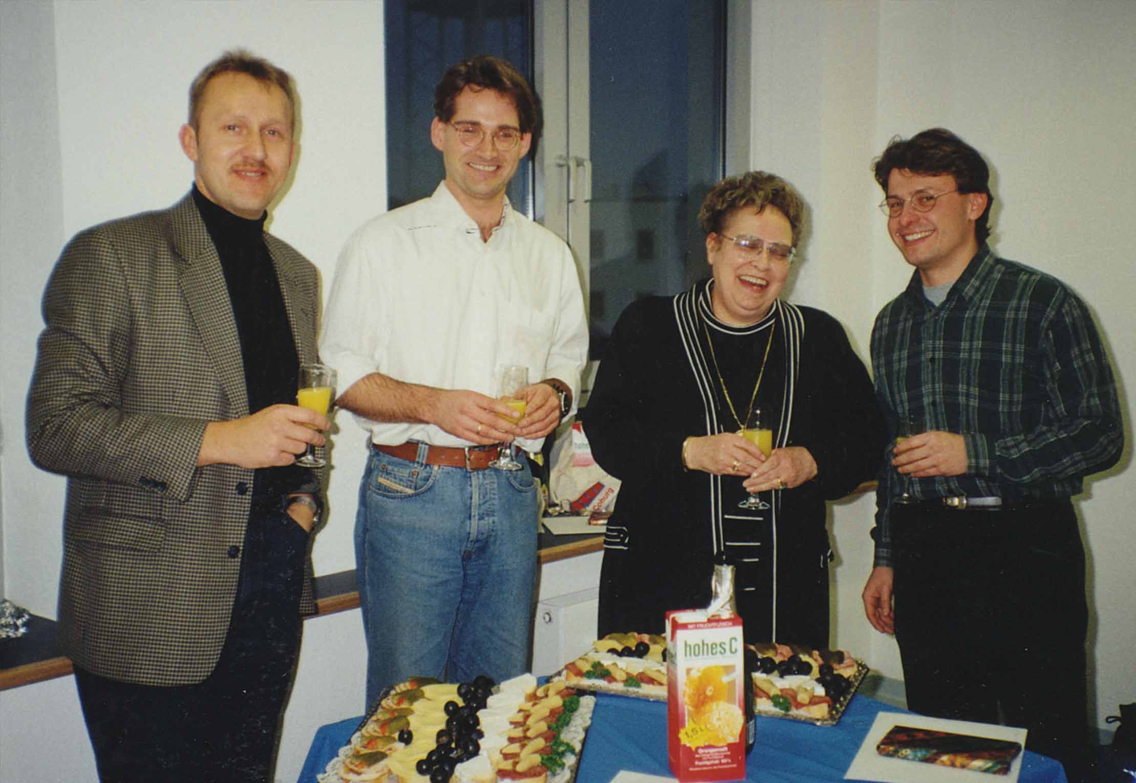 Vier Erwachsene stehen zusammen in einem Innenbereich an einem Tisch mit Essen, Getränken und Saftkartons, lächeln und halten Gläser, als würden sie ein zwangloses Beisammensein oder eine kleine Feier genießen.