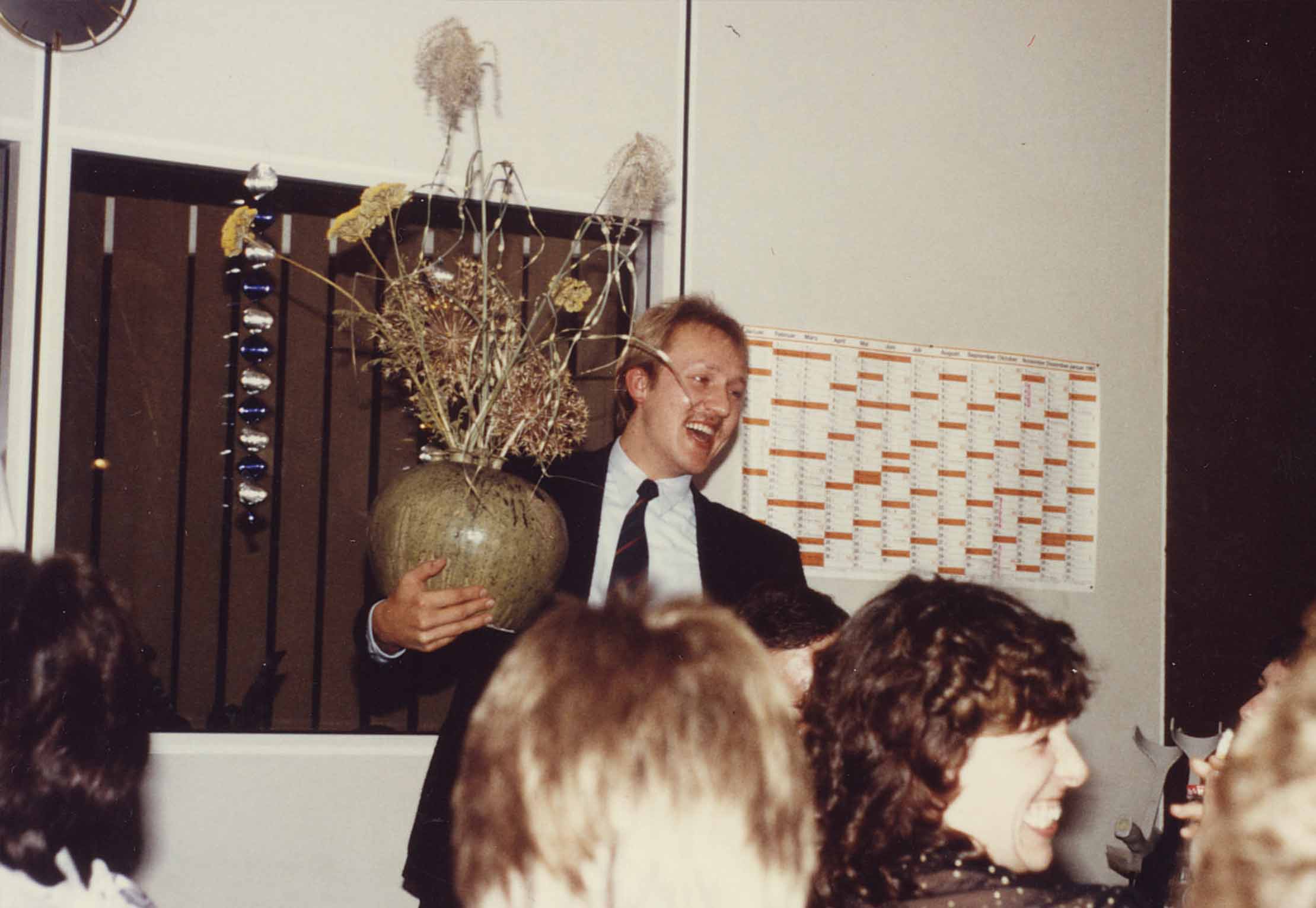 Ein Mann im Anzug hält eine große Vase mit getrockneten Blumen in der Hand und steht lächelnd vor einer Gruppe von Menschen bei einer Versammlung. Im Hintergrund hängen ein Kalender und eine Sonnenbrille an der Wand.