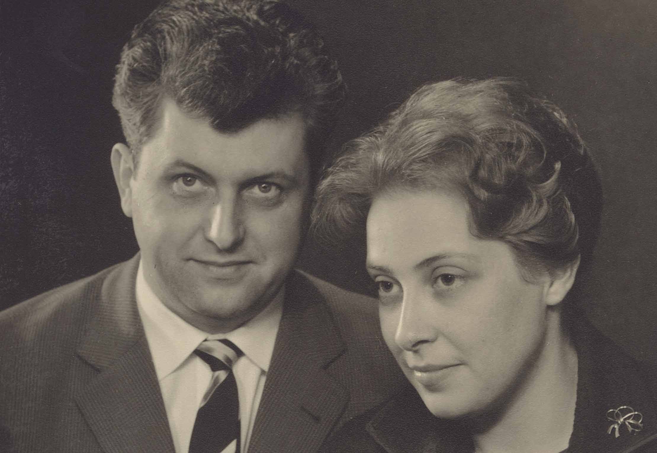 Ein Schwarz-Weiß-Porträt eines Mannes in Anzug und Krawatte neben einer Frau mit kurzen Haaren, die ein dunkles Oberteil mit einer dekorativen Anstecknadel trägt. Beide blicken mit neutralem Gesichtsausdruck leicht vom Bild weg.