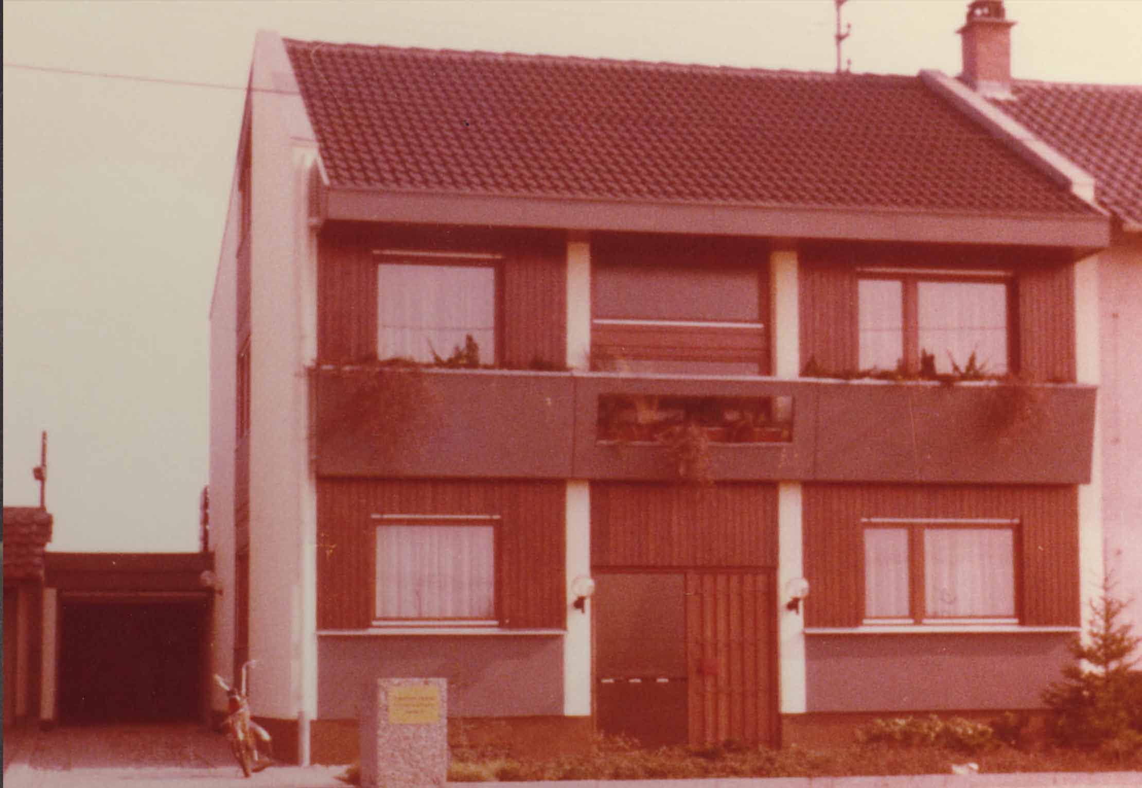 Ein zweistöckiges Haus mit rotem Ziegeldach, braun-beiger Fassade und großen Fenstern. Links neben der Garage parkt ein Fahrrad, vor dem Gebäude steht ein kleiner grüner Busch.