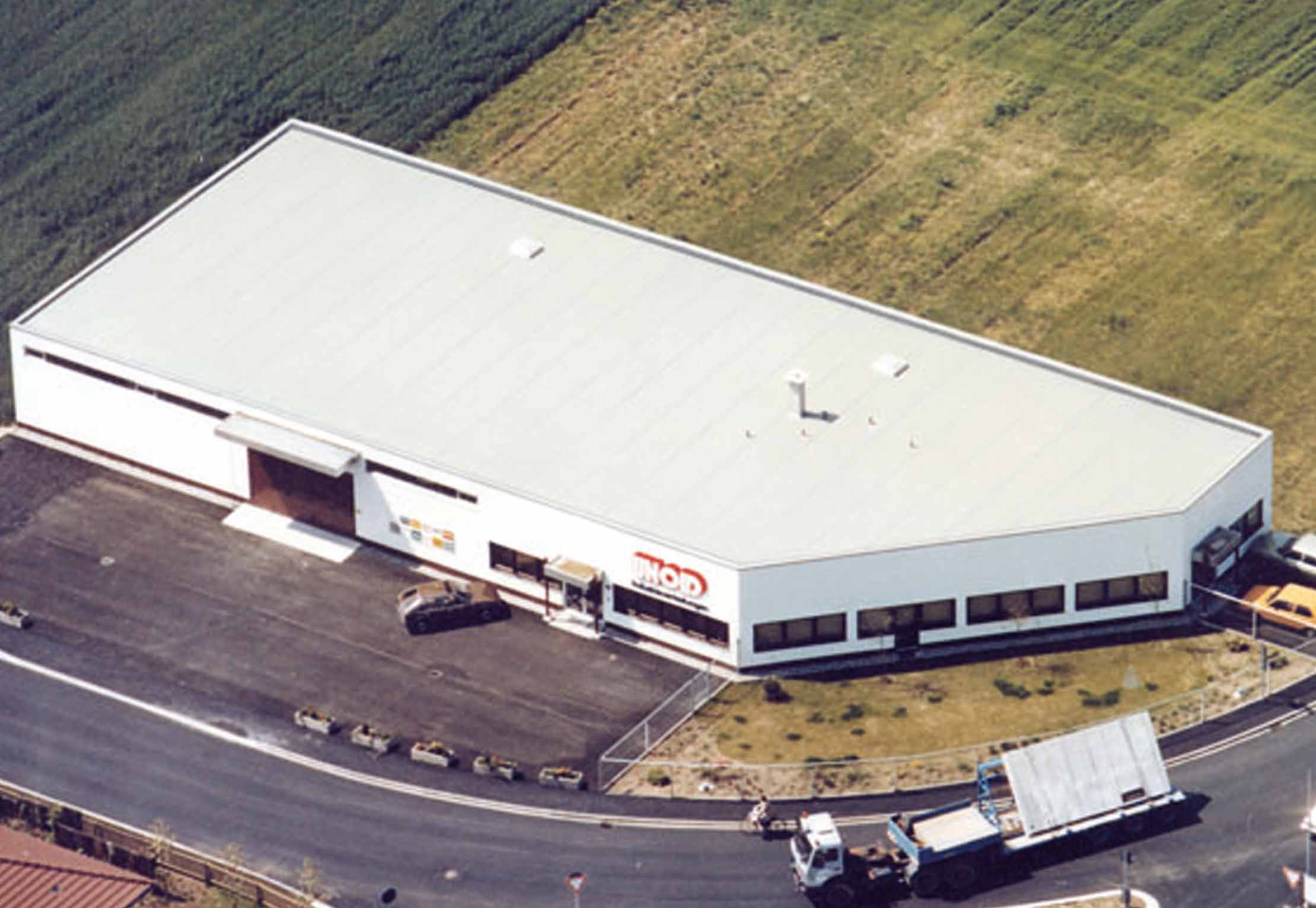 Luftaufnahme eines großen, weißen Industriegebäudes mit Flachdach, umgeben von Gras. Lastwagen und Autos parken auf dem gepflasterten Platz davor, in der Nähe einer Straße, die sich am Gebäude entlangschlängelt.