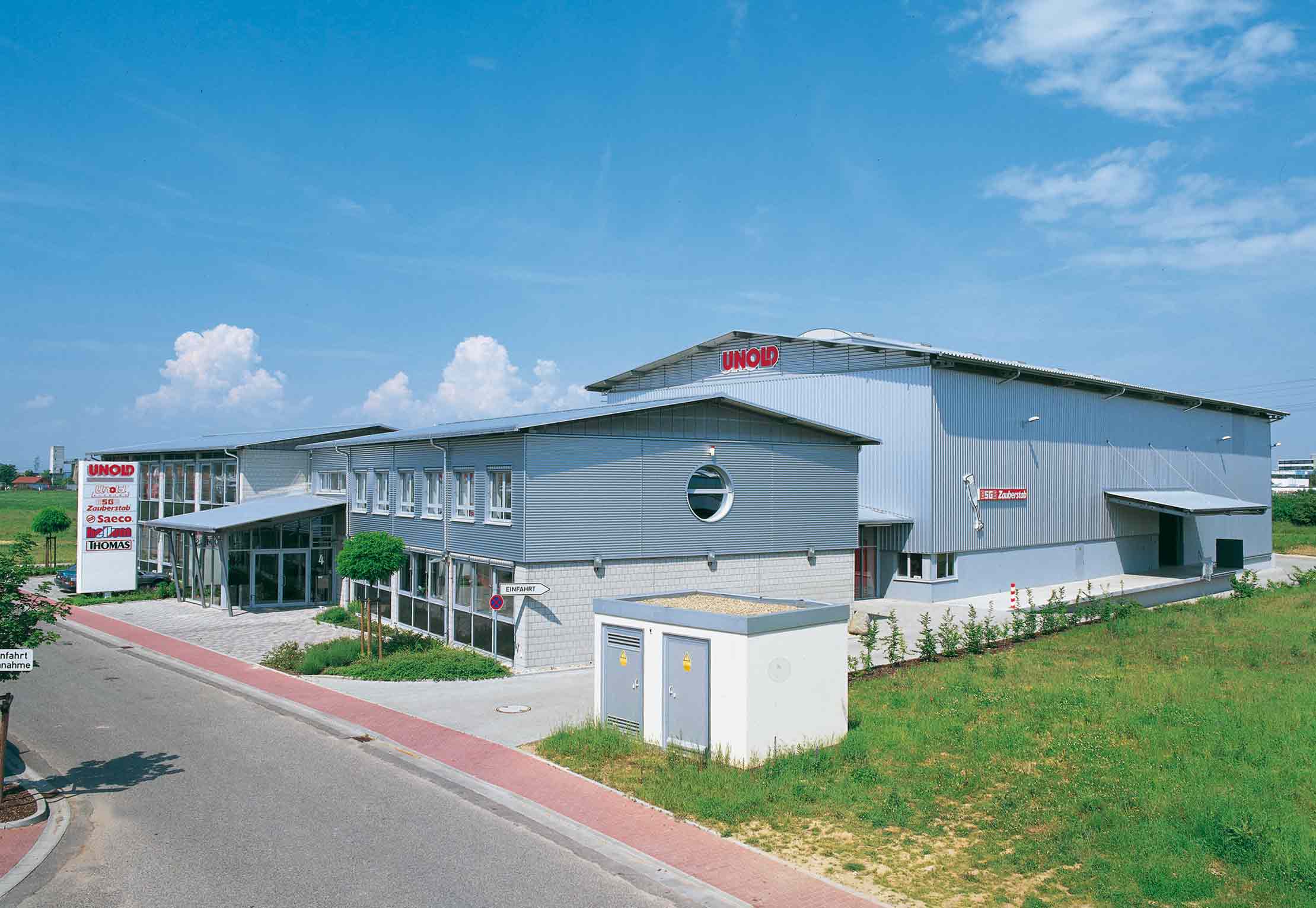 Ein modernes Industriegebäude mit einer grauen Metallfassade, großen Fenstern und roten UNOX-Schildern, neben einer Straße und einer Rasenfläche unter einem blauen Himmel mit einigen Wolken.