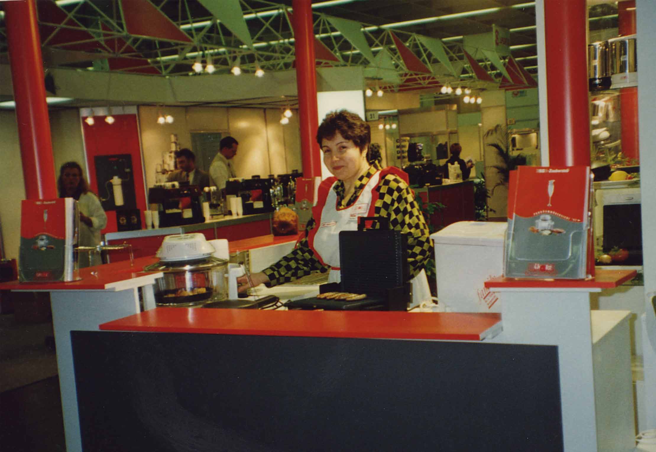 Eine Frau steht auf einer Messe oder Ausstellung hinter einem rot-weißen Demonstrationstresen. Im Hintergrund sind Küchengeräte und Produkte ausgestellt. Die Umgebung ist hell erleuchtet, und im Hintergrund sind mehrere Personen zu sehen.