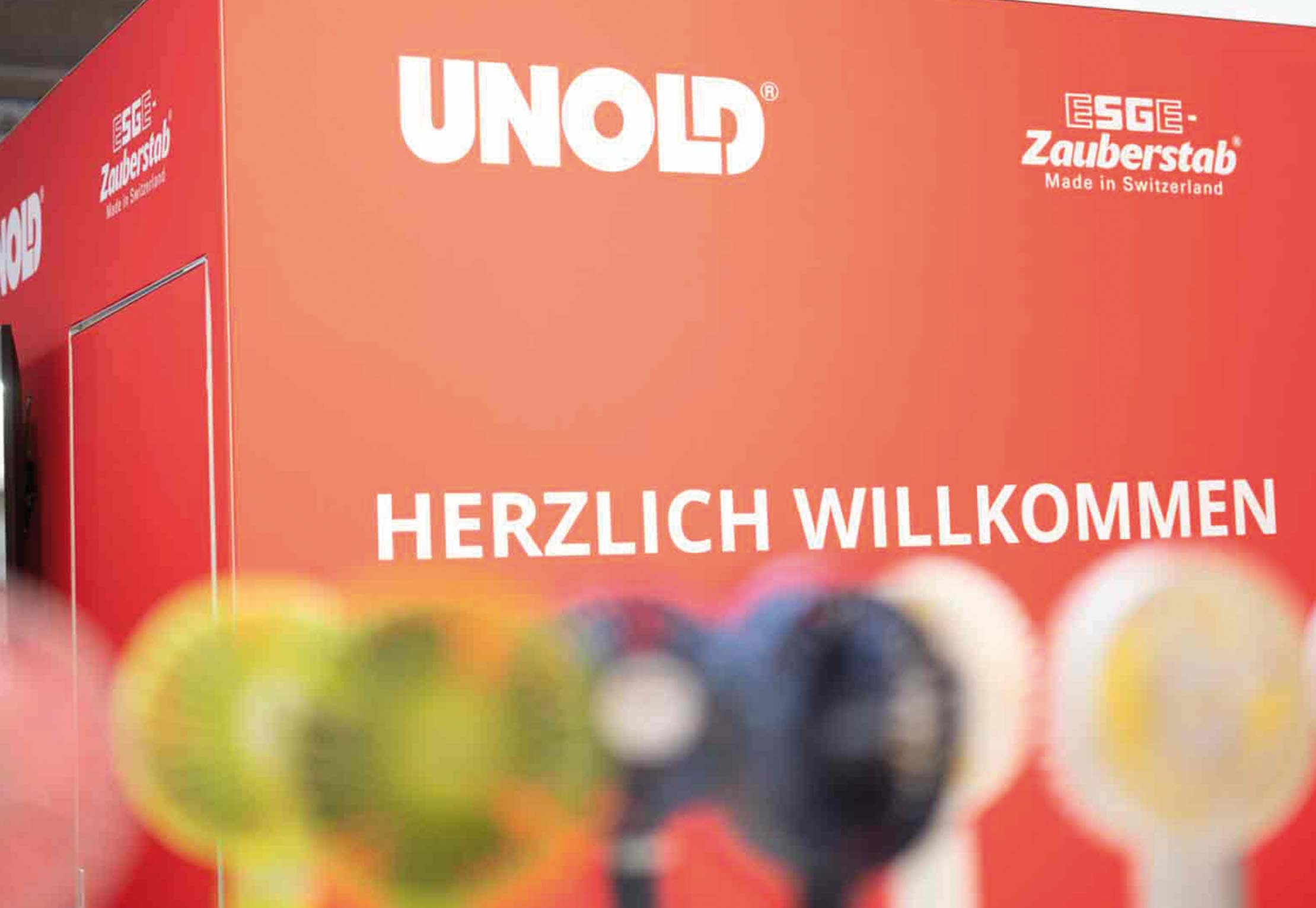 Ein roter Stand zeigt die weißen Schriftzüge „UNOLD“ und „ESGE Zauberstab Made in Switzerland“, darunter „HERZLICH WILLKOMMEN“. Im Vordergrund sind verschwommene bunte Objekte zu sehen.