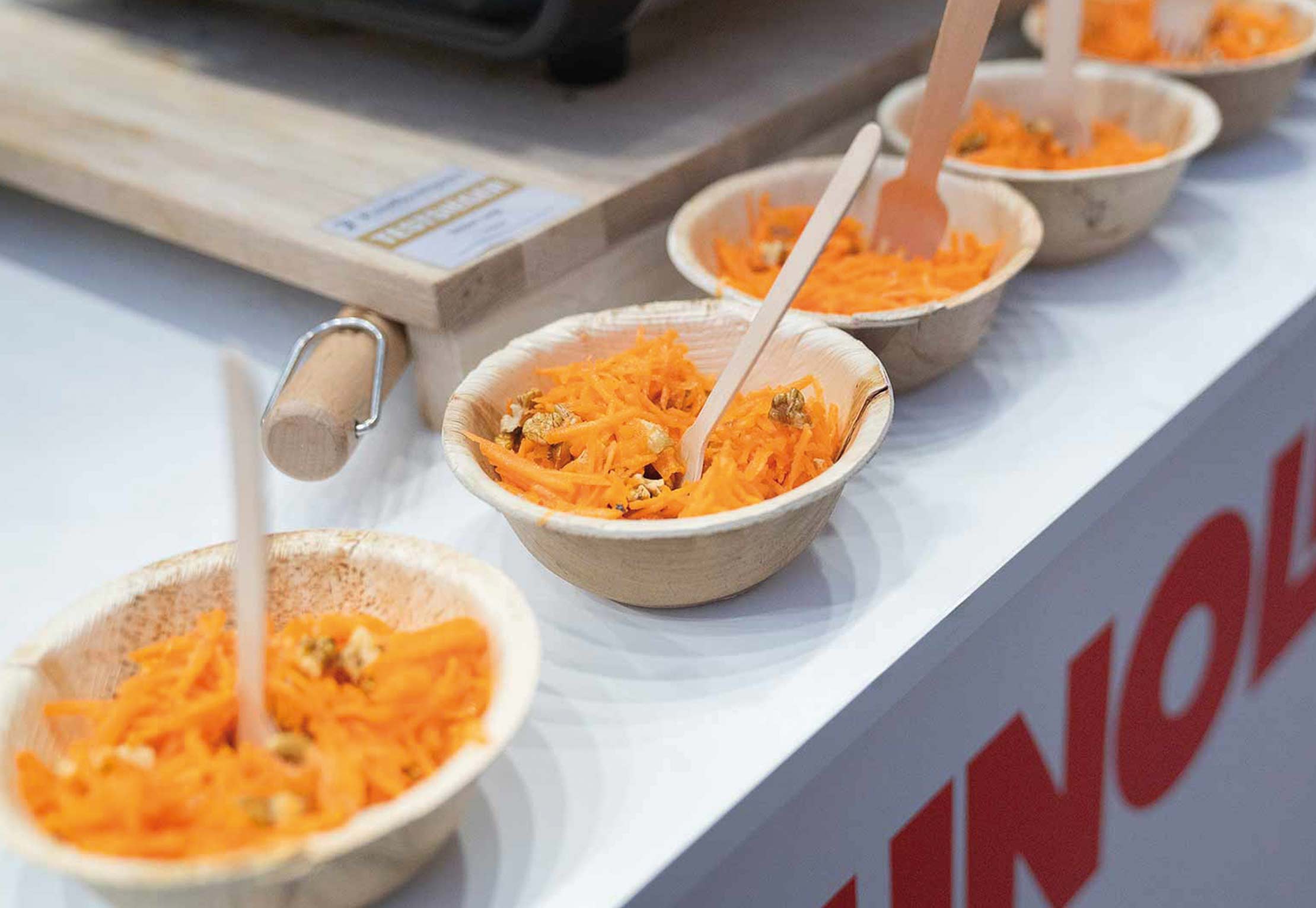 Auf einer Theke stehen mehrere kleine Holzschüsseln, gefüllt mit geraspelten Karotten und Walnüssen, jeweils mit einer Holzgabel, aufgereiht.