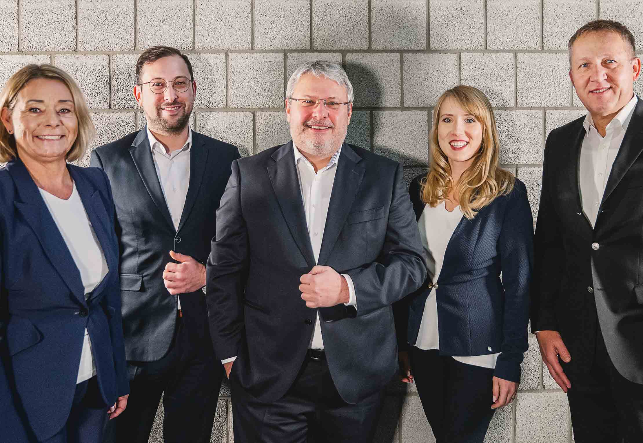 Fünf professionell gekleidete Erwachsene stehen lächelnd in einer Reihe vor einer grauen Betonwand. Drei Männer und zwei Frauen tragen dunkle Anzüge und weiße Hemden und wirken selbstbewusst und zugänglich.